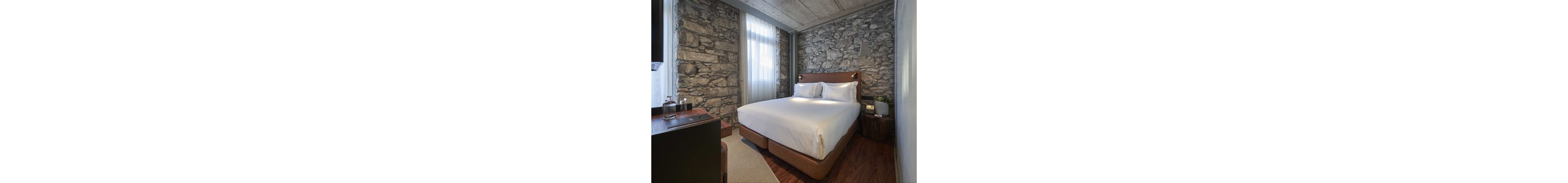 A neatly made double bed with white linens sits centered in a compact hotel room. The space features exposed stone walls, a concrete ceiling, and sheer curtains allowing soft daylight to enter. A small wooden nightstand, contemporary pendant light, and polished wood floor add warm accents to the minimalist decor. On a side table, two glass water bottles and tumblers suggest guest amenities in this comfortable urban retreat.