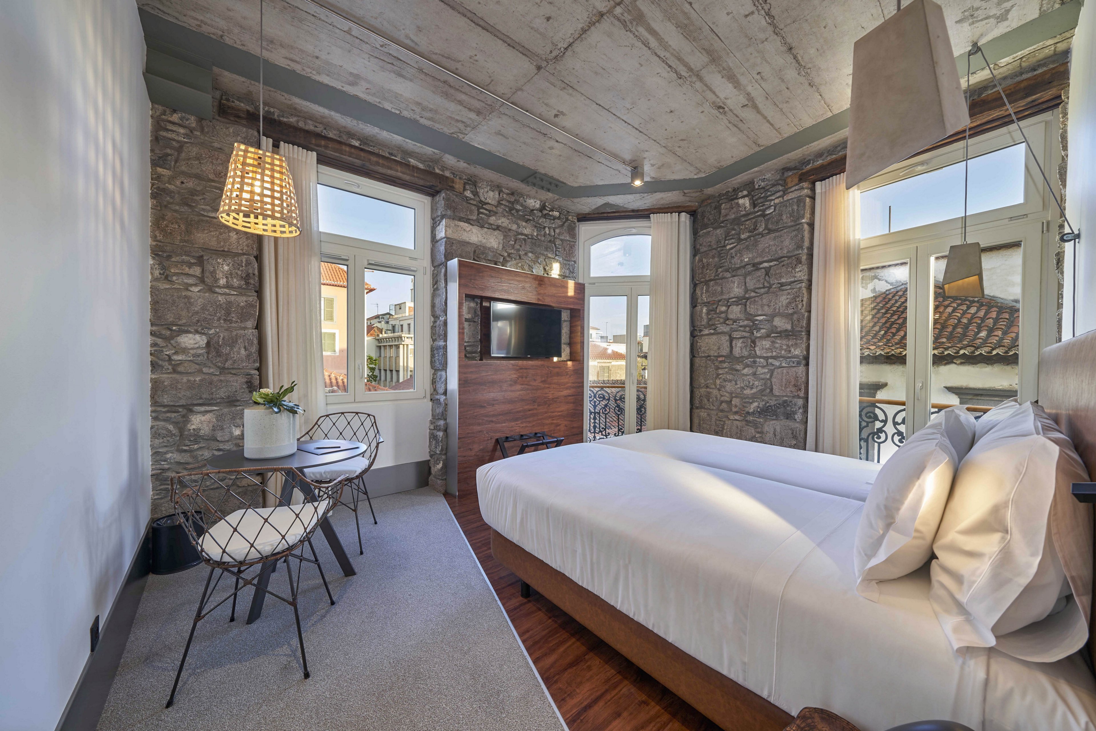 A contemporary hotel room features a neatly made double bed facing a wall-mounted television. Exposed stone walls and large windows bring in natural light and views of surrounding rooftops. A small seating area with wire-frame chairs and a round table sits beside a potted plant, adding a touch of greenery. Industrial-style ceiling and pendant lighting complement the modern, minimalist decor.