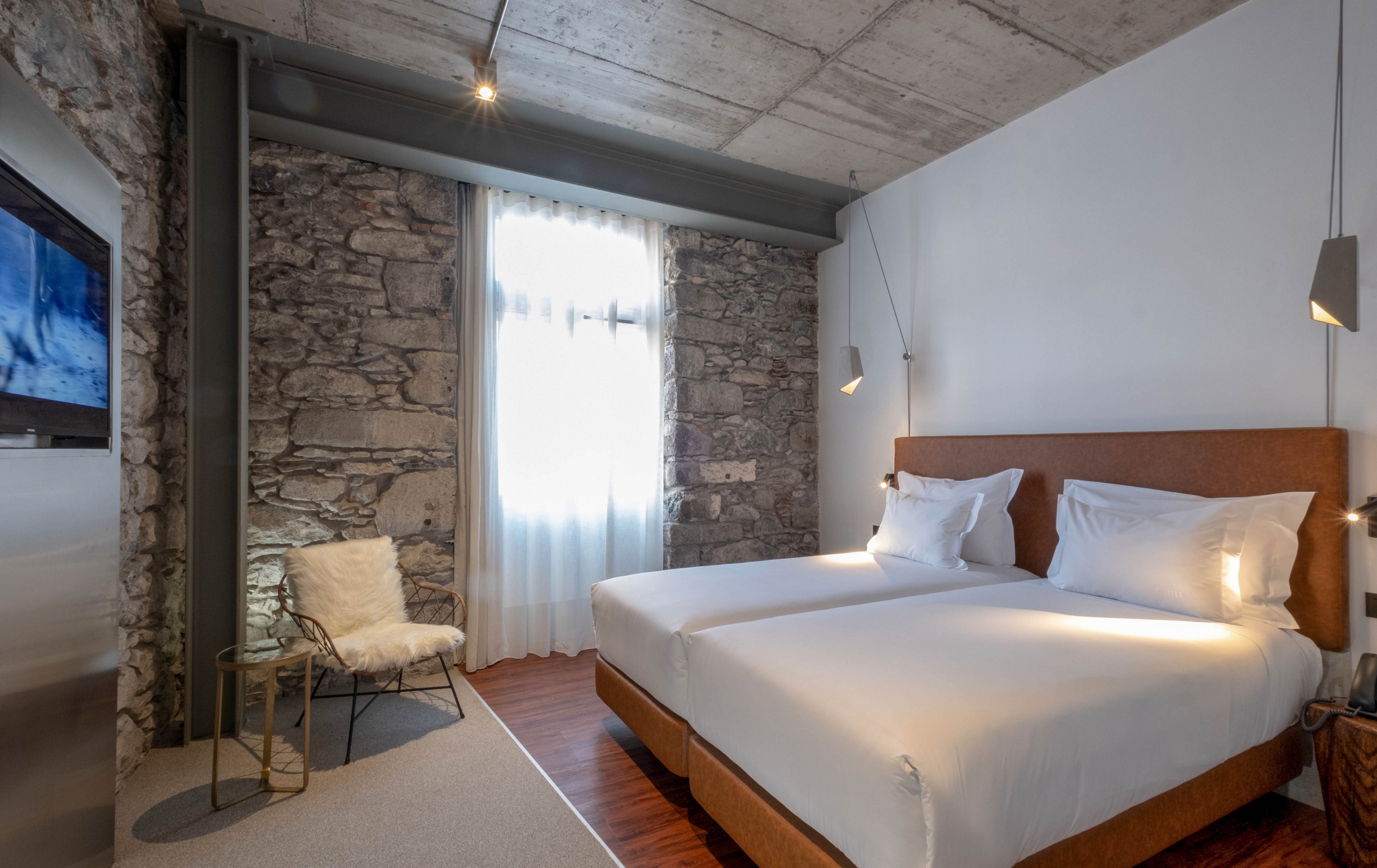 Contemporary hotel bedroom featuring two neatly made twin beds with white linens and wooden bases. The space combines a rustic stone wall with smooth white surfaces and an exposed concrete ceiling for an industrial-chic look. A window with sheer curtains lets in natural light, while minimalist pendant lights and a small chair with a fur throw add cozy accents. Warm wood flooring and simple decor create a clean, inviting atmosphere.