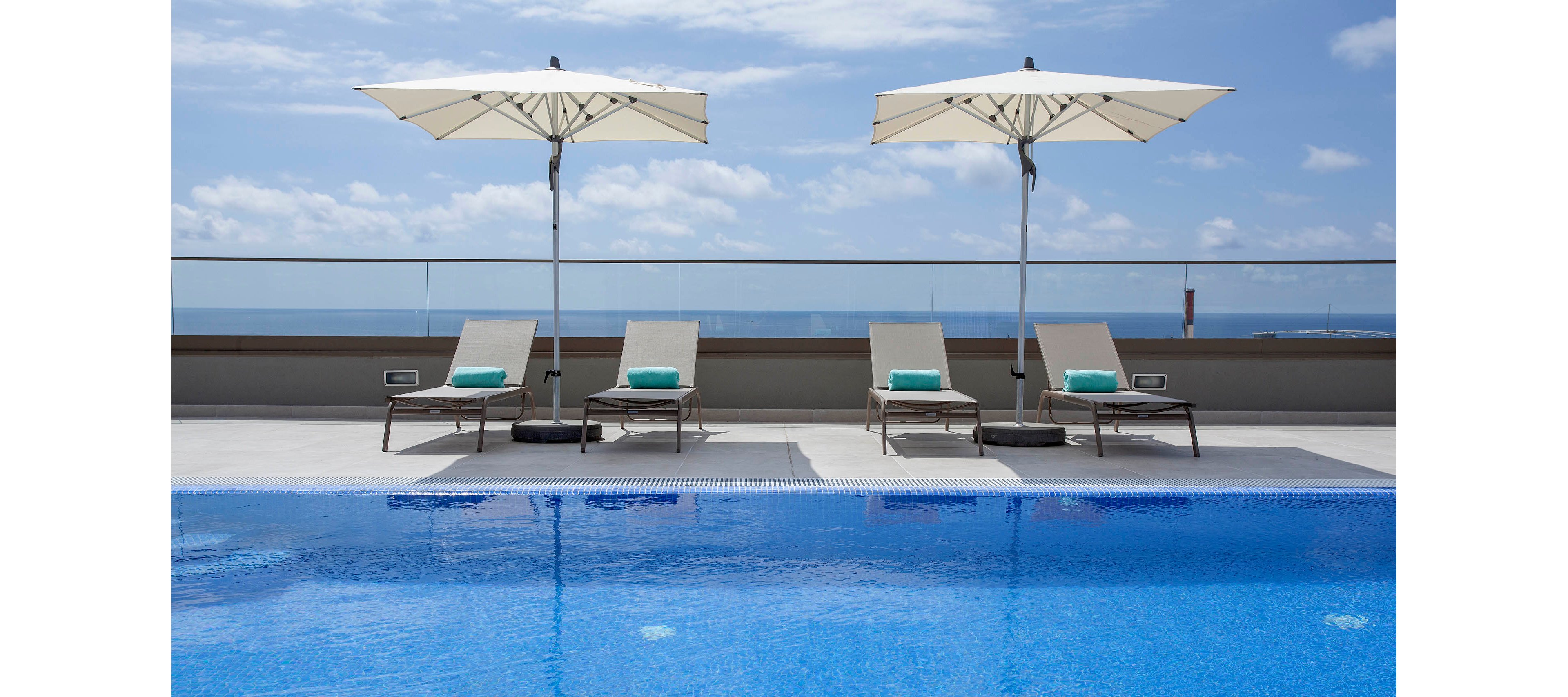 A serene rooftop pool area features four beige lounge chairs with turquoise towels, shaded by two large white umbrellas. The setting overlooks a clear blue sky and glass railing, creating a tranquil and inviting atmosphere. The pool water is vibrant blue, reflecting the sky above, and the overall mood is calm and relaxing. No people are present, emphasizing a peaceful, resort-like ambiance.