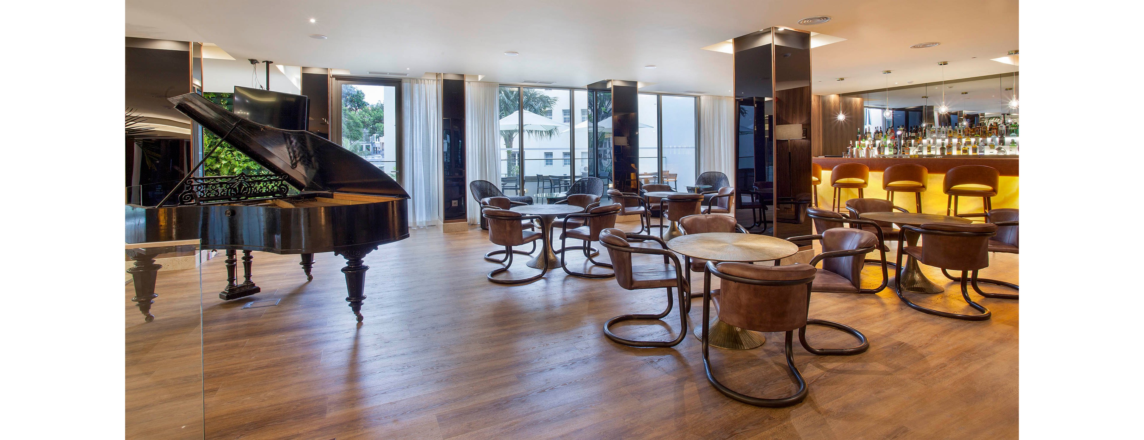 A spacious, contemporary hotel lounge features a glossy black grand piano as a focal point. The setting includes round wooden tables with matching chairs, polished wood flooring, and a well-lit bar with high stools. Large windows allow natural light to fill the room, highlighting the clean and elegant interior design. The atmosphere is sophisticated and inviting, ideal for social gatherings or relaxing.