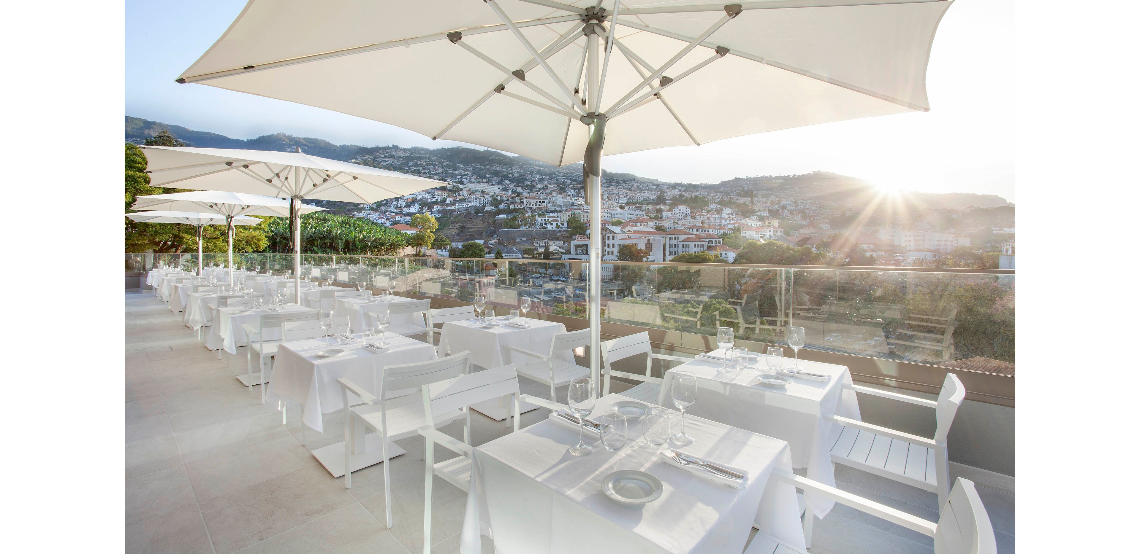A modern outdoor restaurant terrace features neatly arranged white tables and chairs under large umbrellas. The setting overlooks a scenic cityscape with hills in the background, bathed in soft sunlight. Table settings include glassware and cutlery, creating a refined and inviting atmosphere. The overall mood is serene and sophisticated, ideal for upscale dining experiences.