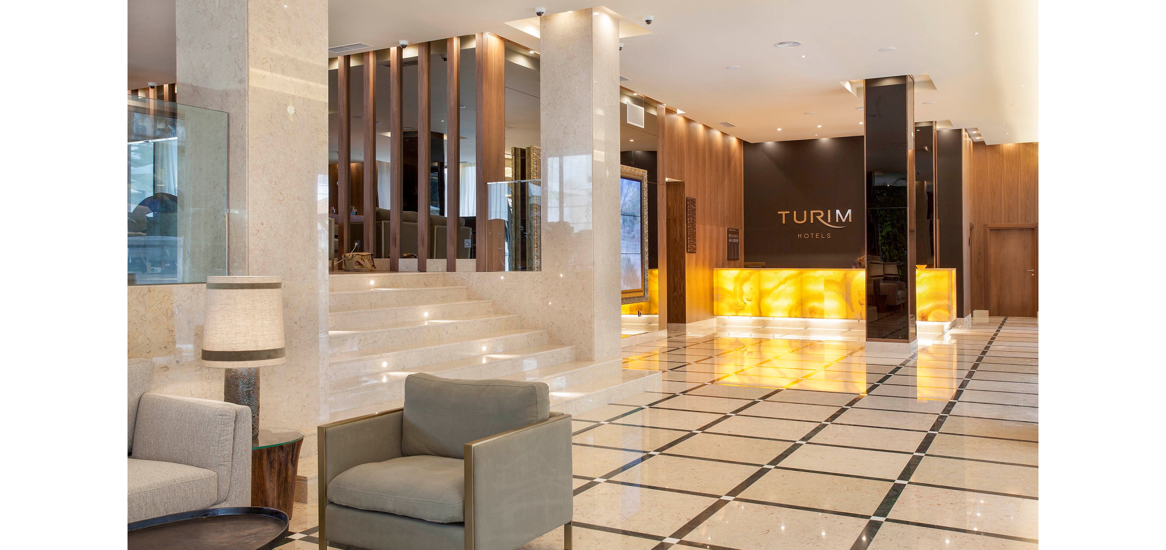 The image shows the lobby of a Turim hotel, featuring a sleek and modern interior design. The setting includes polished marble floors, a glowing yellow reception desk, and comfortable seating areas. Warm lighting and neutral tones create a welcoming and upscale atmosphere. The word 'TURIM' is clearly visible on the back wall, indicating the hotel name.