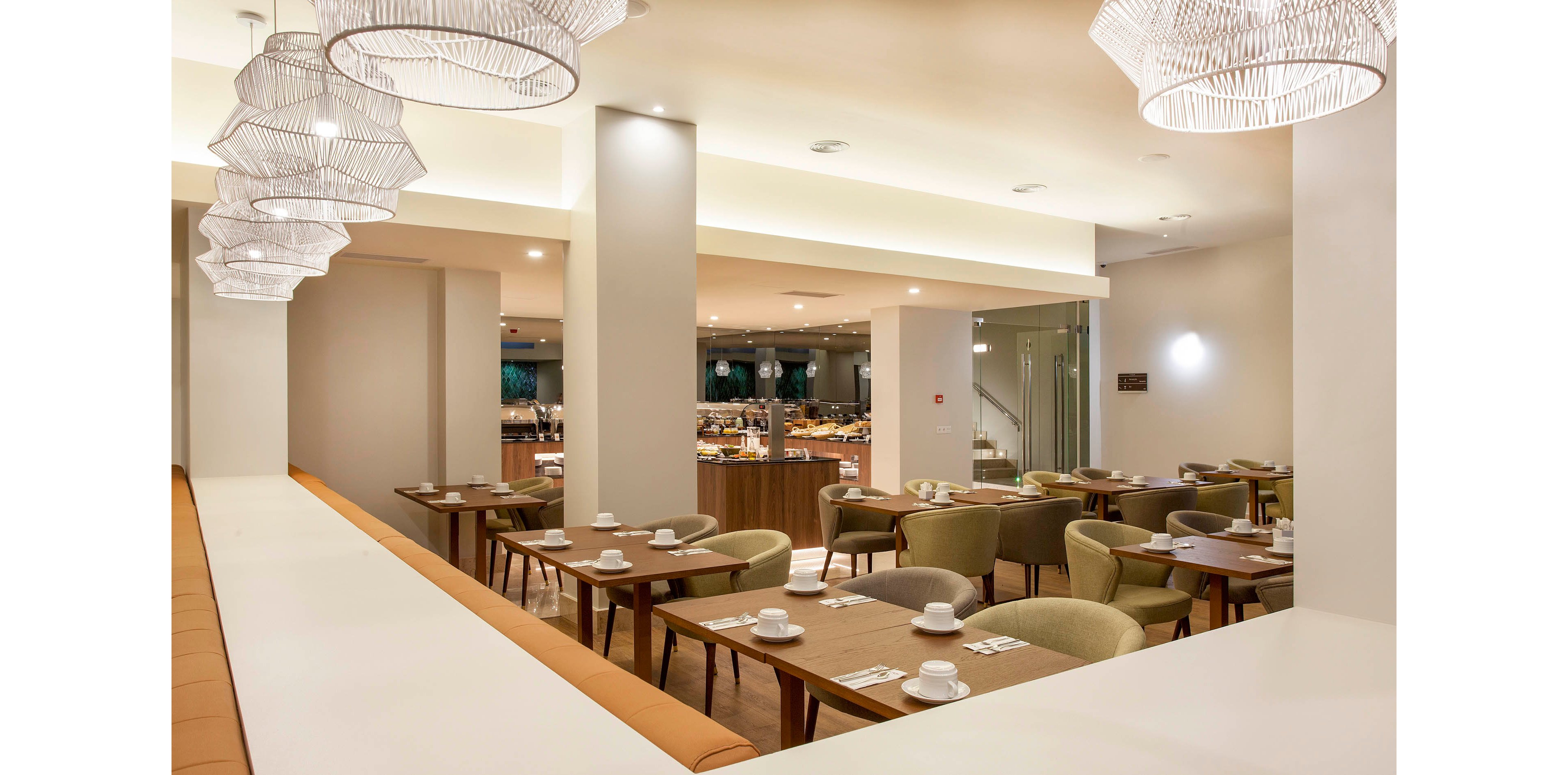 A contemporary restaurant interior features neatly arranged tables and chairs, each set with white cups and saucers. The setting is well-lit with stylish pendant lights and a visible buffet area in the background. The color palette is neutral with warm wood tones and soft green seating. The atmosphere is calm and inviting, suitable for casual or business dining. No visible text or numbers are present in the image.