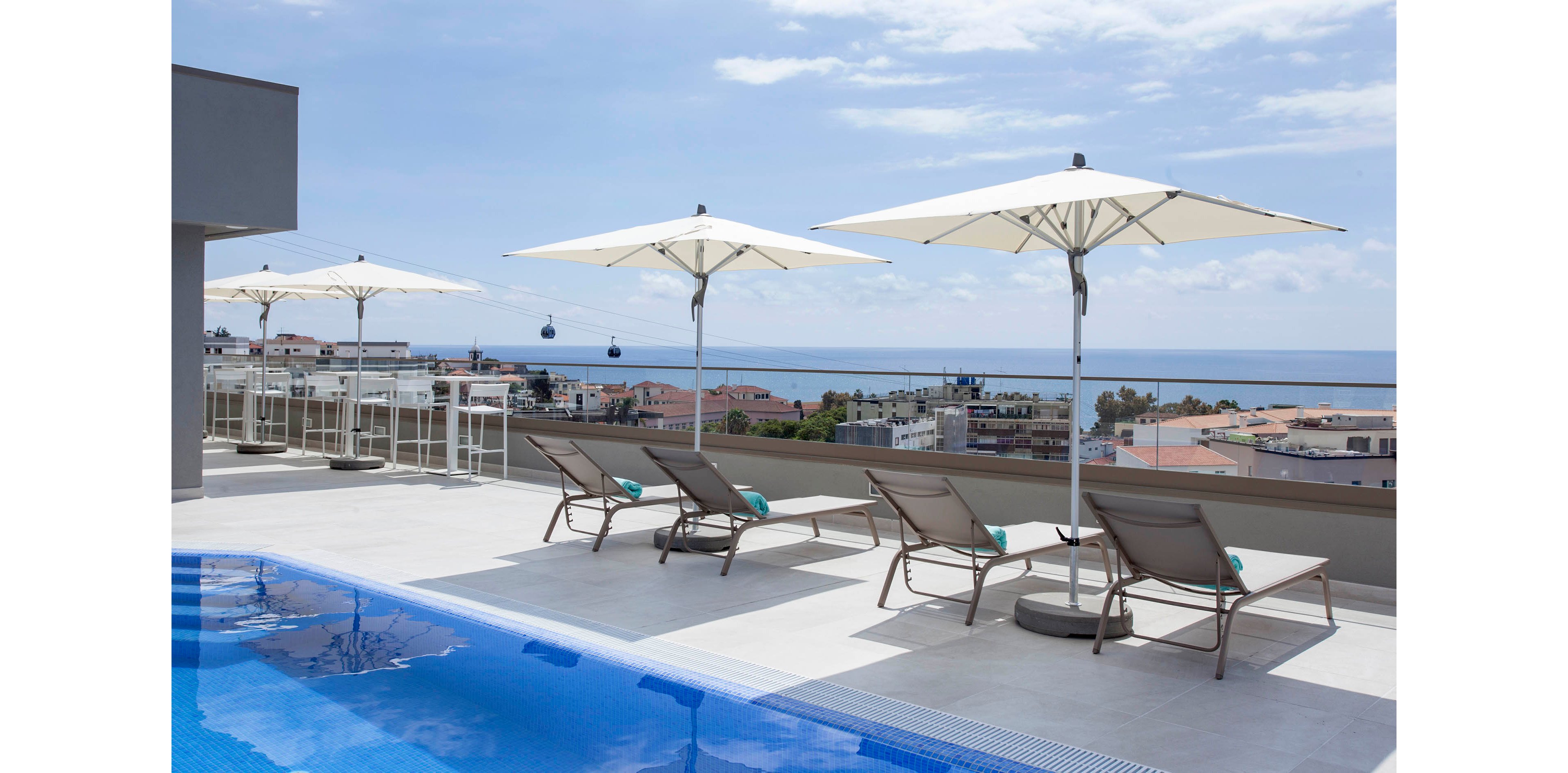 A contemporary rooftop terrace features a clear blue swimming pool, several reclining loungers, and large white umbrellas. The setting overlooks a coastal cityscape with buildings and the ocean visible in the background. The scene is bright and airy, evoking a relaxed and luxurious mood, with clean lines and minimalistic outdoor furniture.