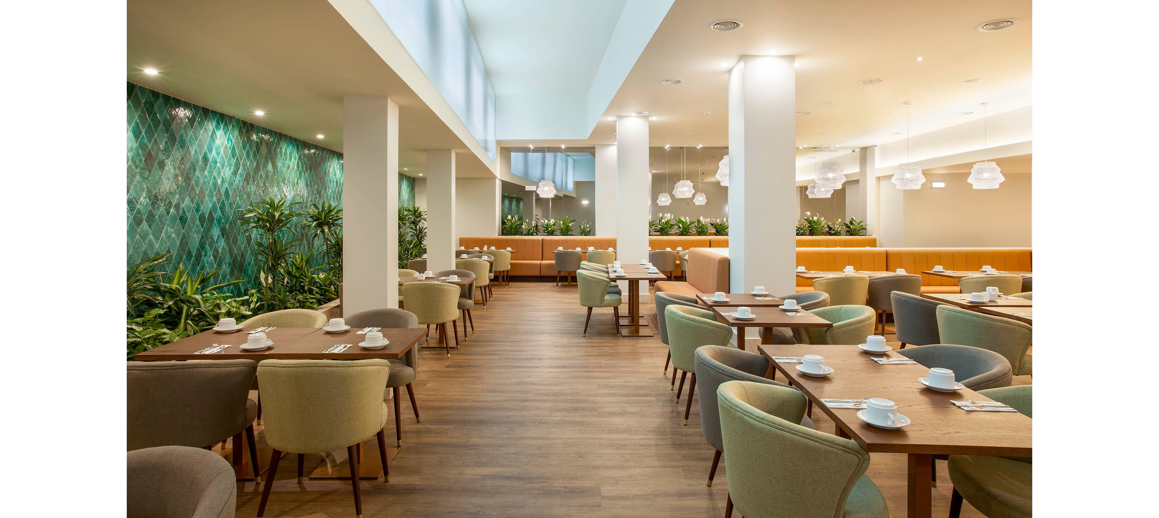 Spacious restaurant dining area featuring contemporary furniture and a striking green tiled accent wall. Tables are set with white dishware and cups, and the space is filled with natural light from large windows. The setting is calm and inviting, with soft earth tones and indoor plants enhancing the relaxed atmosphere. No visible people or text are present in the image.