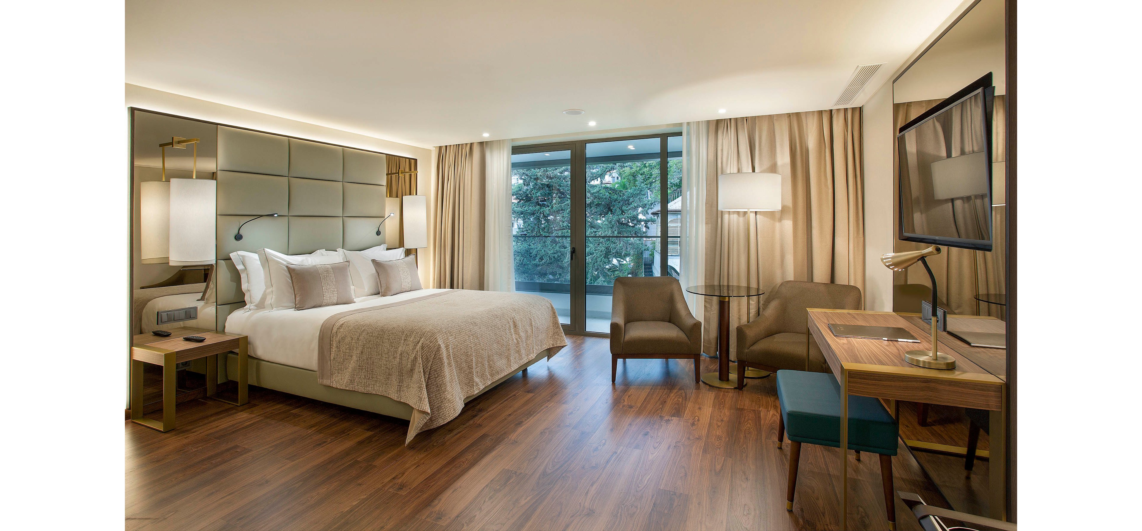 A spacious, contemporary hotel room features a large double bed with neutral bedding, two armchairs, and a work desk. Floor-to-ceiling windows open to a balcony with a view of trees, allowing natural light to fill the space. The room is decorated in warm beige and brown tones, with wood flooring and soft ambient lighting creating a calm, inviting atmosphere. No visible text or numbers are present in the image.