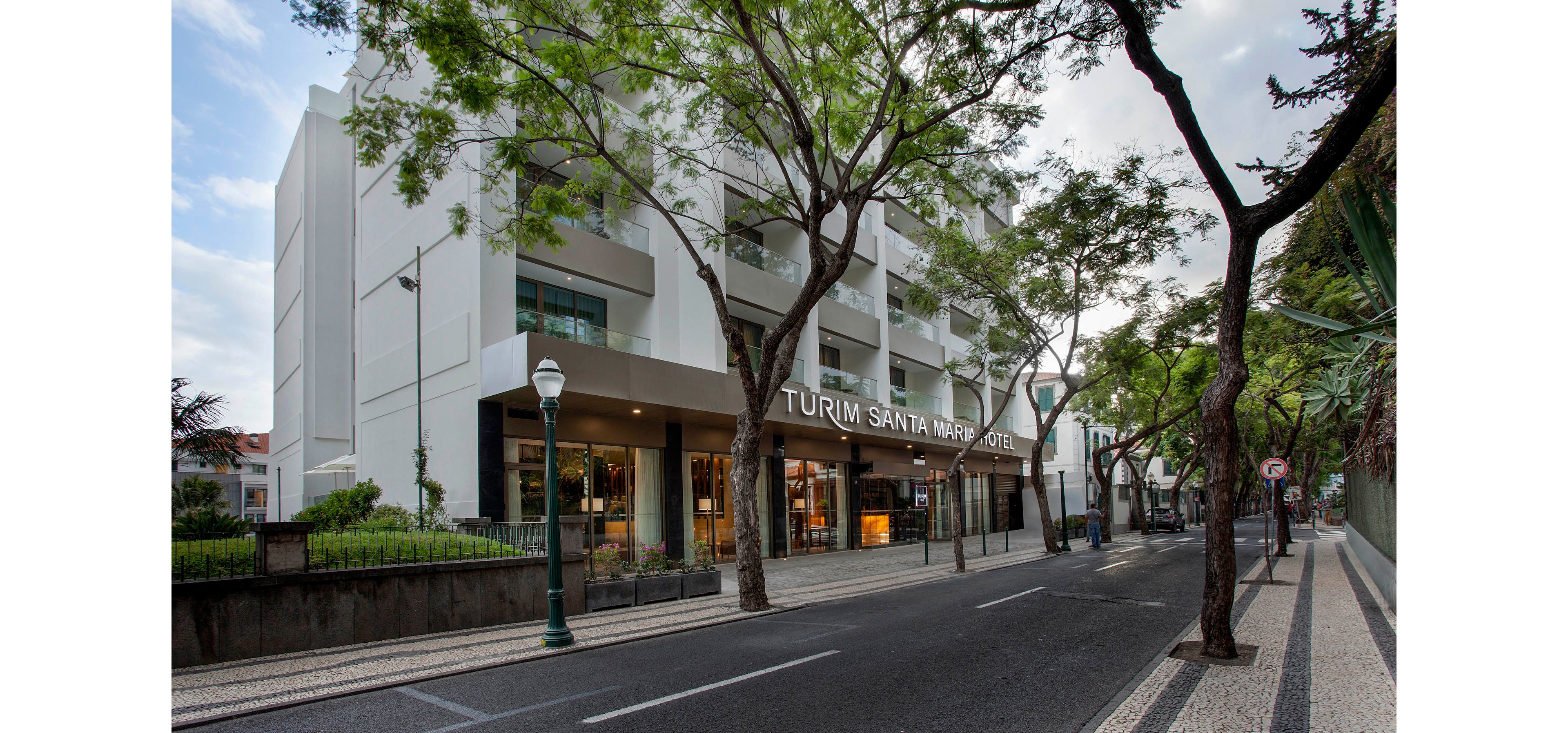 The image shows the modern exterior of the Turim Santa Maria Hotel, located along a tree-lined urban street. The hotel facade is white with large windows, and the entrance is clearly visible with the name displayed above. The street is empty, featuring patterned sidewalks, street lamps, and lush green trees, creating a calm and inviting atmosphere.