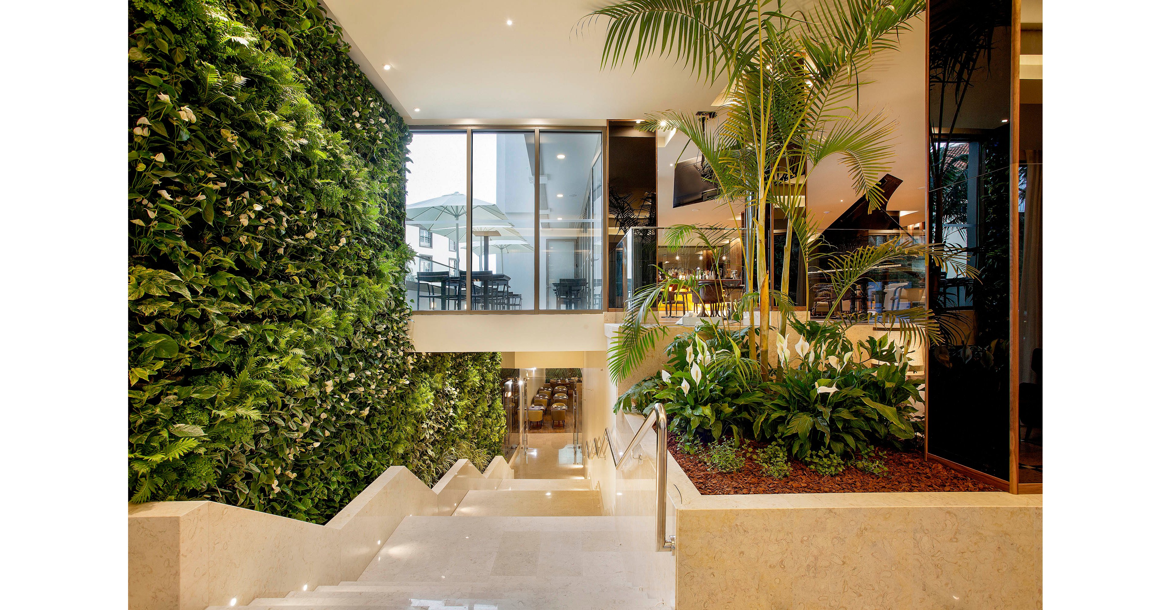 A contemporary indoor staircase features a striking vertical garden wall filled with green foliage. The setting is bright and airy, with large windows allowing natural light to flood the space. Polished stone steps and indoor tropical plants enhance the fresh, inviting atmosphere. The overall mood is modern and tranquil, with a focus on biophilic design elements.