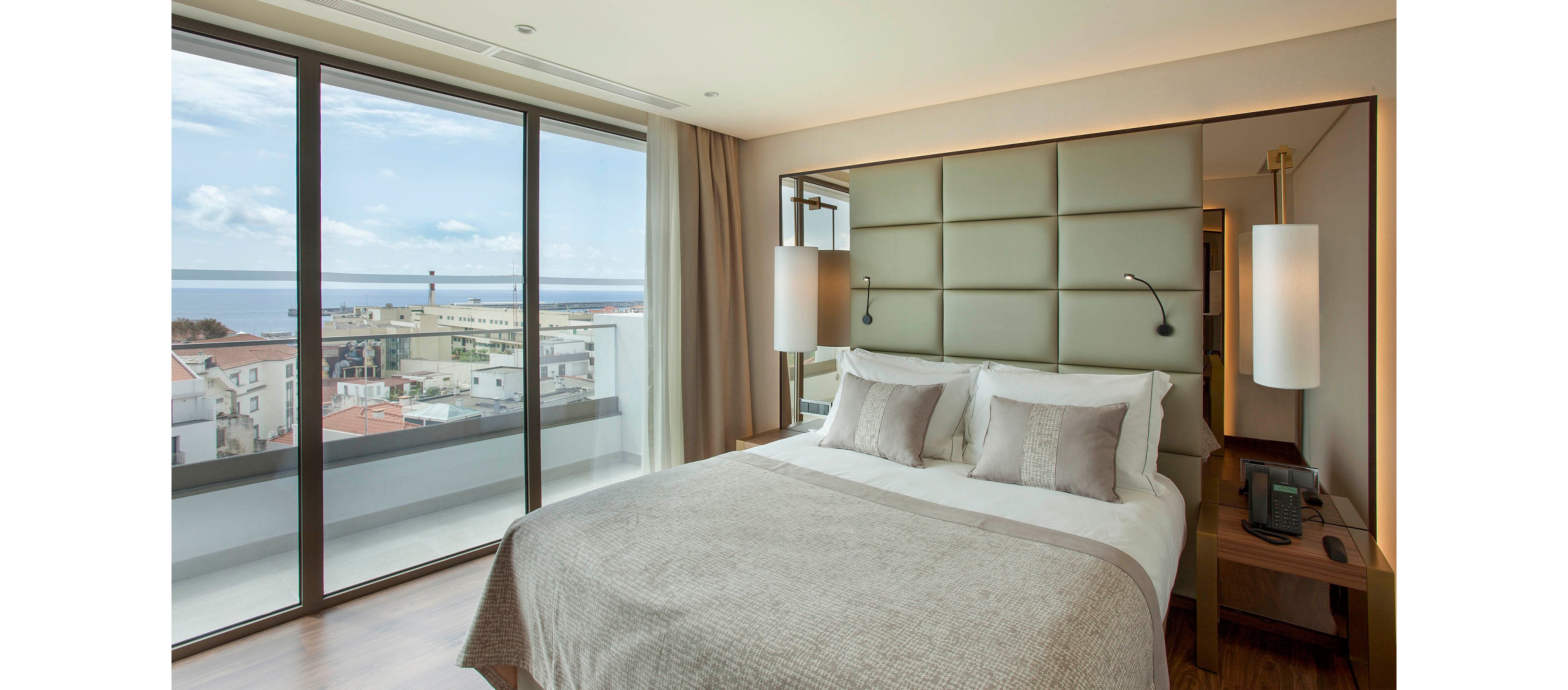 A contemporary hotel room features a large bed with neutral bedding and a padded headboard. Floor-to-ceiling windows offer a panoramic view of the city and ocean beyond. The room is decorated in soft beige and gray tones, with minimalist bedside tables and pendant lighting. The atmosphere is calm and inviting, ideal for relaxation or business travel.