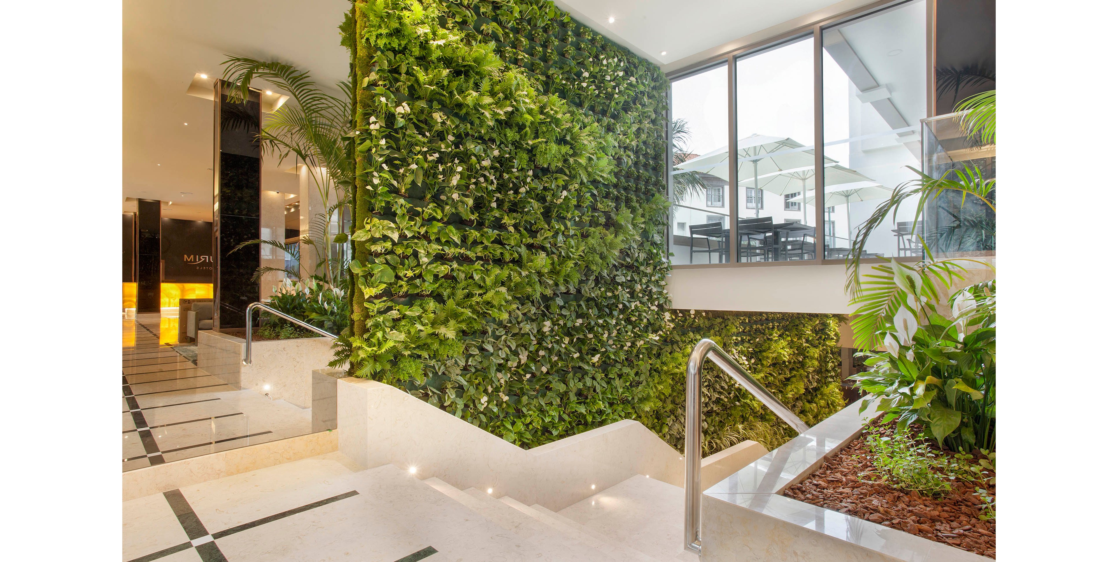 A contemporary indoor lobby features a striking vertical garden wall filled with various green plants. The space is bright and airy, with large windows allowing natural light to flood the area. Sleek marble stairs and stainless steel railings add a modern touch, while the greenery creates a refreshing, eco-friendly atmosphere. The overall palette is neutral with vibrant green accents.