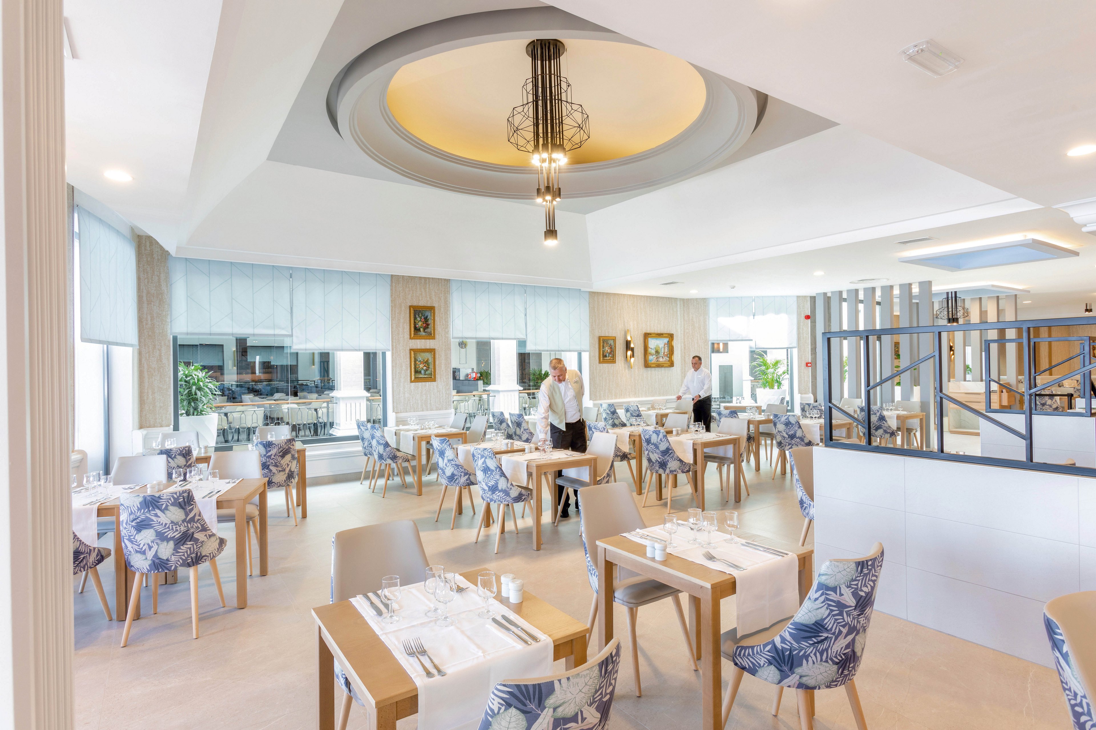 The image shows a bright, contemporary restaurant with neatly arranged tables and chairs featuring blue and white patterned upholstery. A female staff member is seen standing in the dining area, which is decorated with framed artwork and large windows letting in natural light. The setting is clean and inviting, with neutral tones and stylish lighting fixtures contributing to a relaxed atmosphere. No visible numeric values or identifiable brands are present.