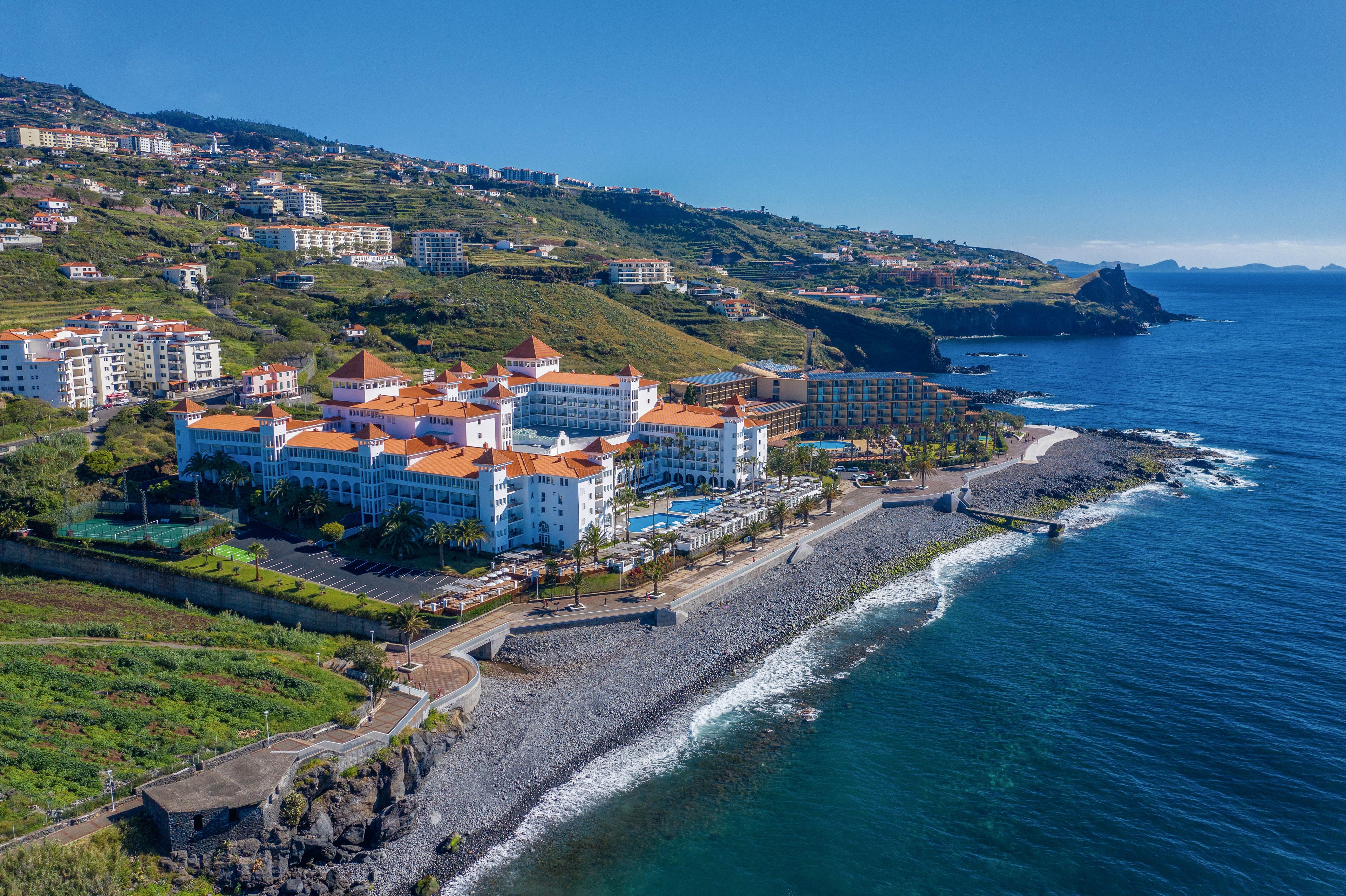 A large seaside resort complex with distinctive red rooftops sits along a rocky shoreline, surrounded by lush green hills and residential buildings. The setting is a sunny day with clear blue skies and calm ocean waves gently reaching the shore. The scene captures the vibrant contrast between the white buildings, orange roofs, and deep blue sea, evoking a relaxed and inviting mood.