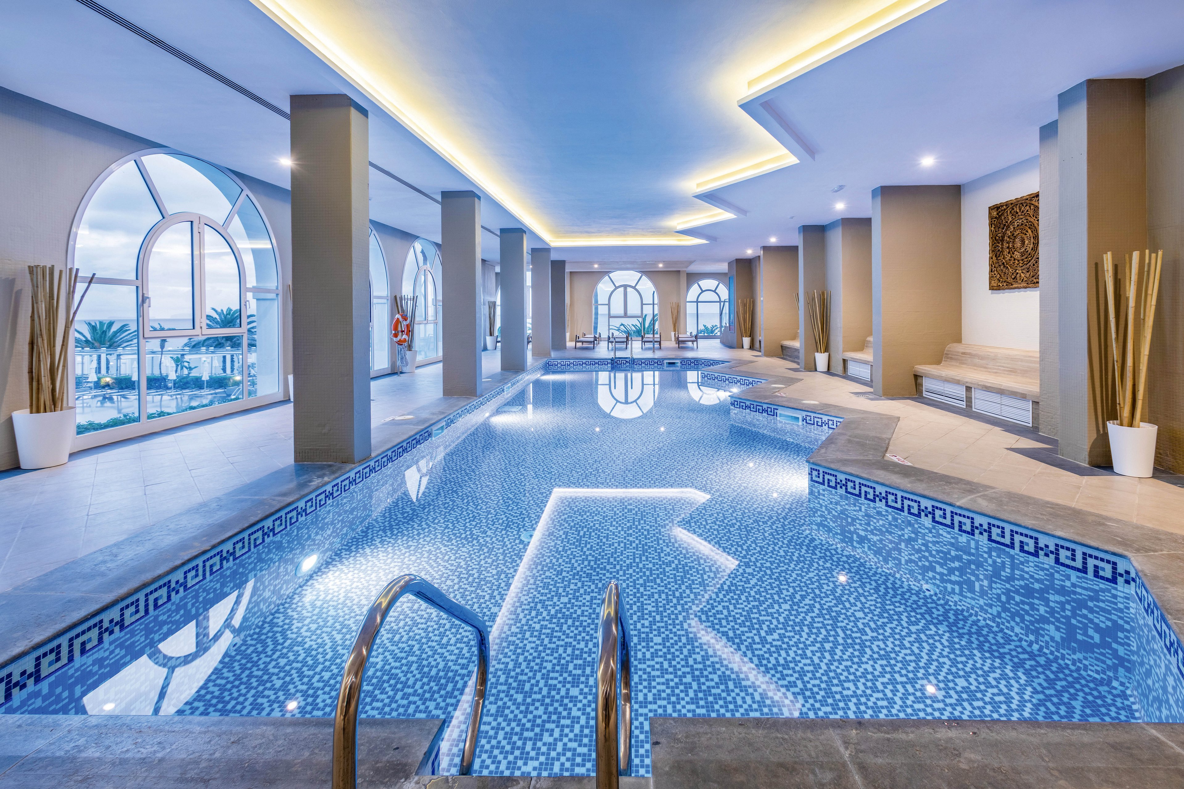 A spacious indoor swimming pool is featured in a contemporary setting with large arched windows allowing natural light to flood the area. The pool is surrounded by sleek columns, decorative vases, and comfortable seating, creating a relaxing atmosphere. The mosaic tile pattern in the pool and the soft blue palette contribute to a tranquil mood. No visible text or numeric values are present in the image.
