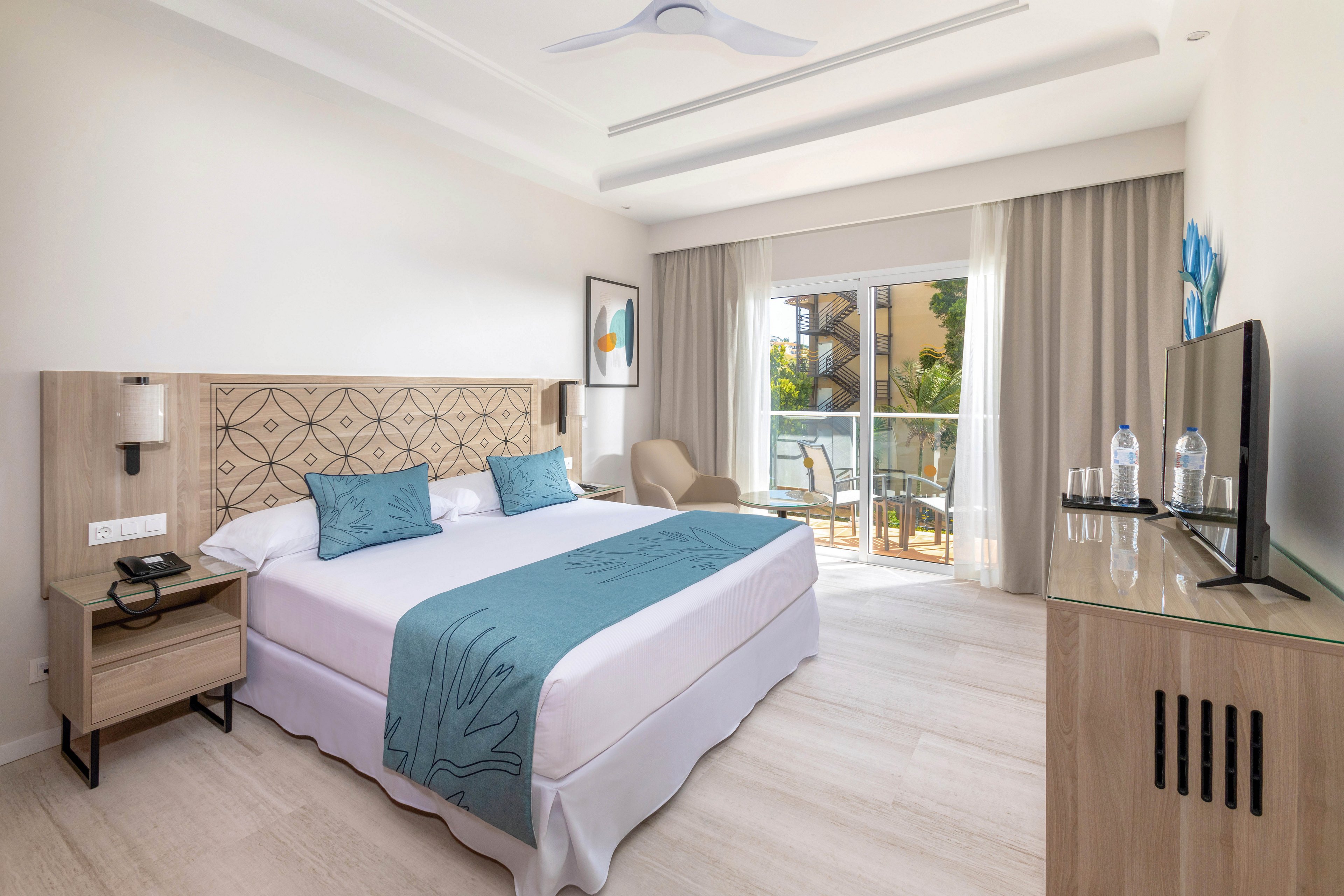 A contemporary hotel room features a double bed with blue pillows and a matching runner, set against a decorative headboard. The space includes a flat-screen TV, two bottles of water, a phone, and a comfortable chair near the window. Natural light fills the room, highlighting the neutral palette and inviting balcony view.
