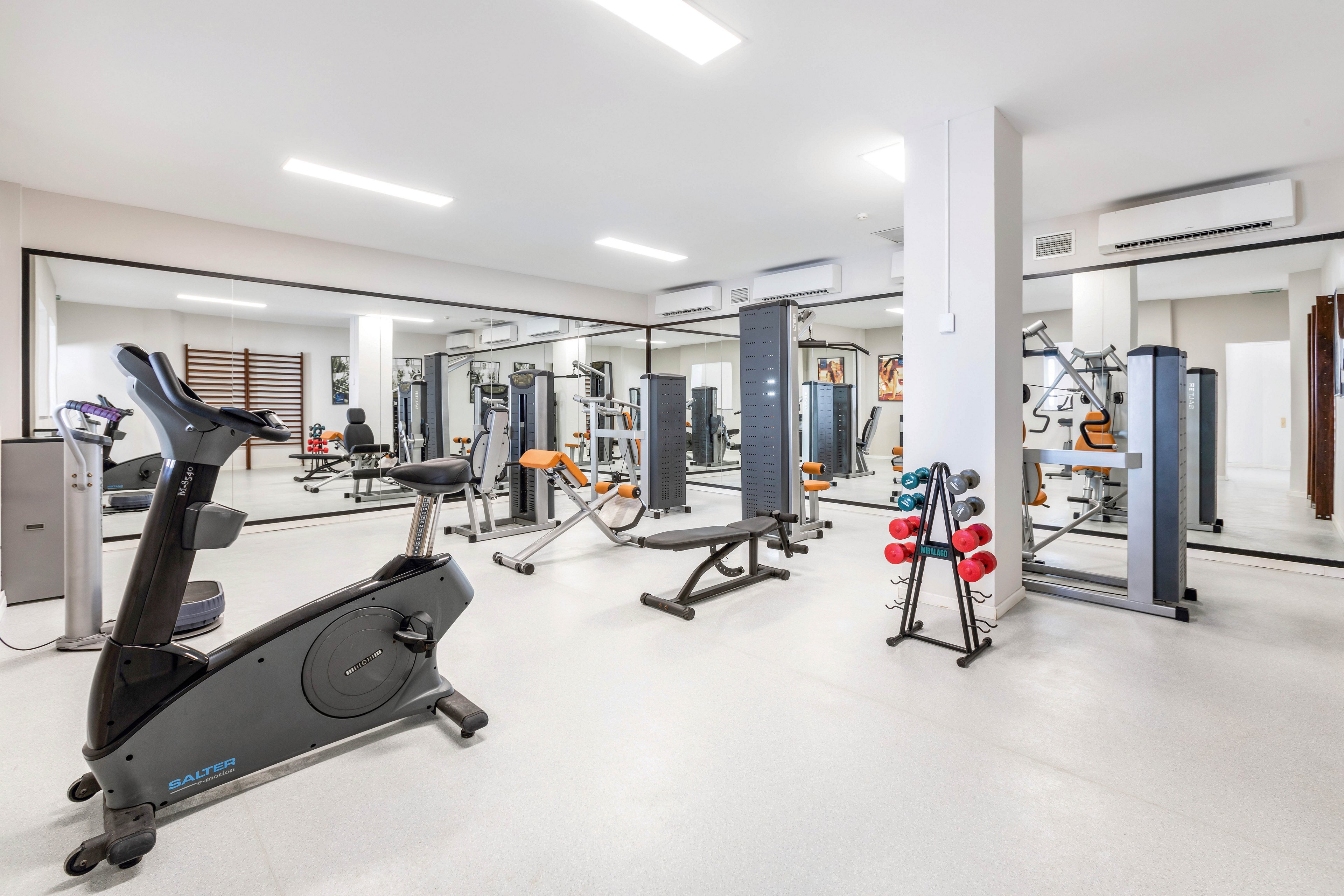 A spacious, well-lit gym features various exercise machines including a stationary bike, weight bench, and strength training equipment. The room has large wall mirrors, white floors, and a rack of red dumbbells. The setting is clean and organized, with a bright, neutral color palette and no people present.