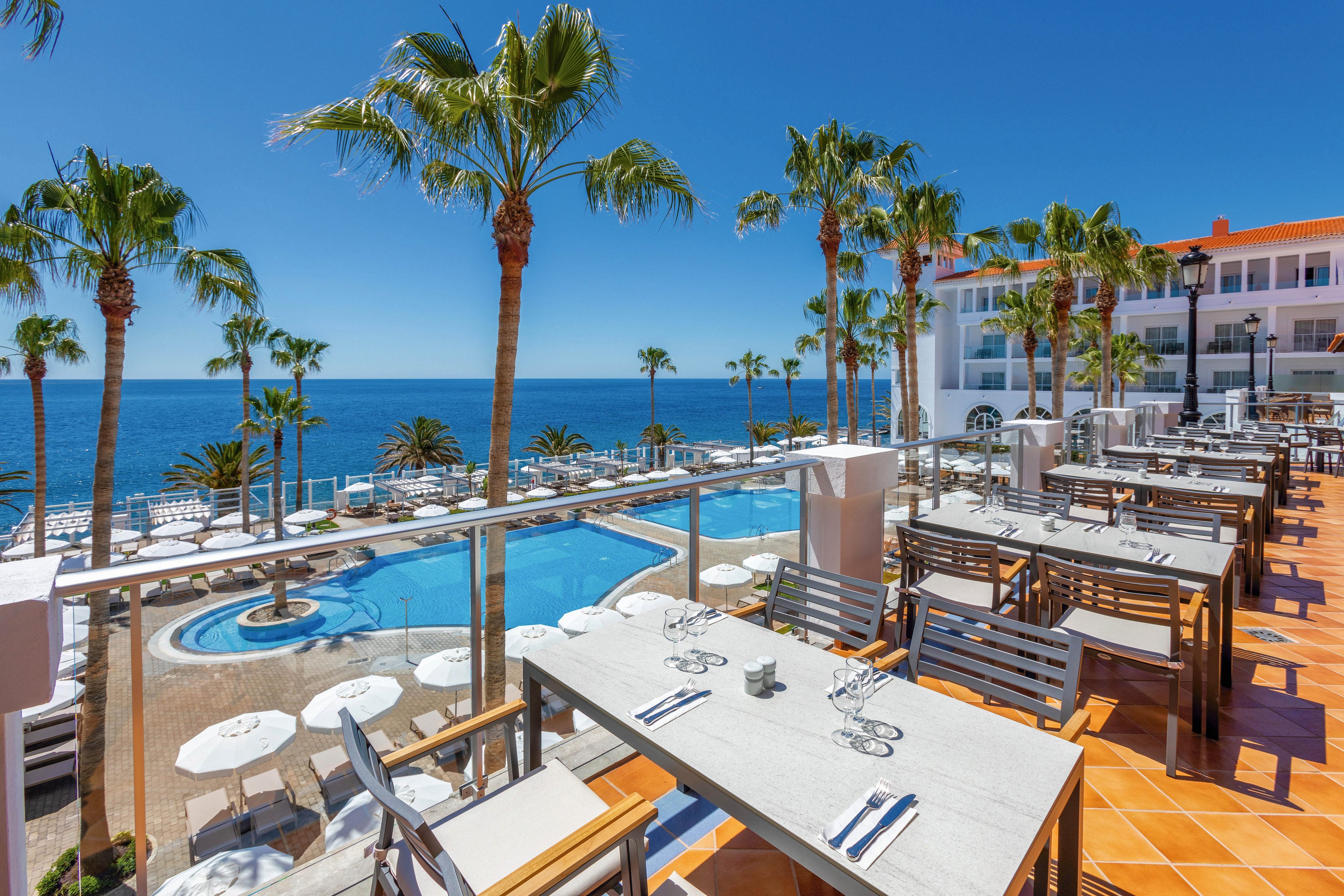 The image features a spacious outdoor dining area with neatly arranged tables and chairs, set on a terrace with a stunning view of a swimming pool and the ocean beyond. Palm trees and white umbrellas add a tropical ambiance, while the clear blue sky and bright sunlight create a vibrant, inviting atmosphere. The setting appears to be part of a modern hotel or resort, with clean lines and contemporary decor.