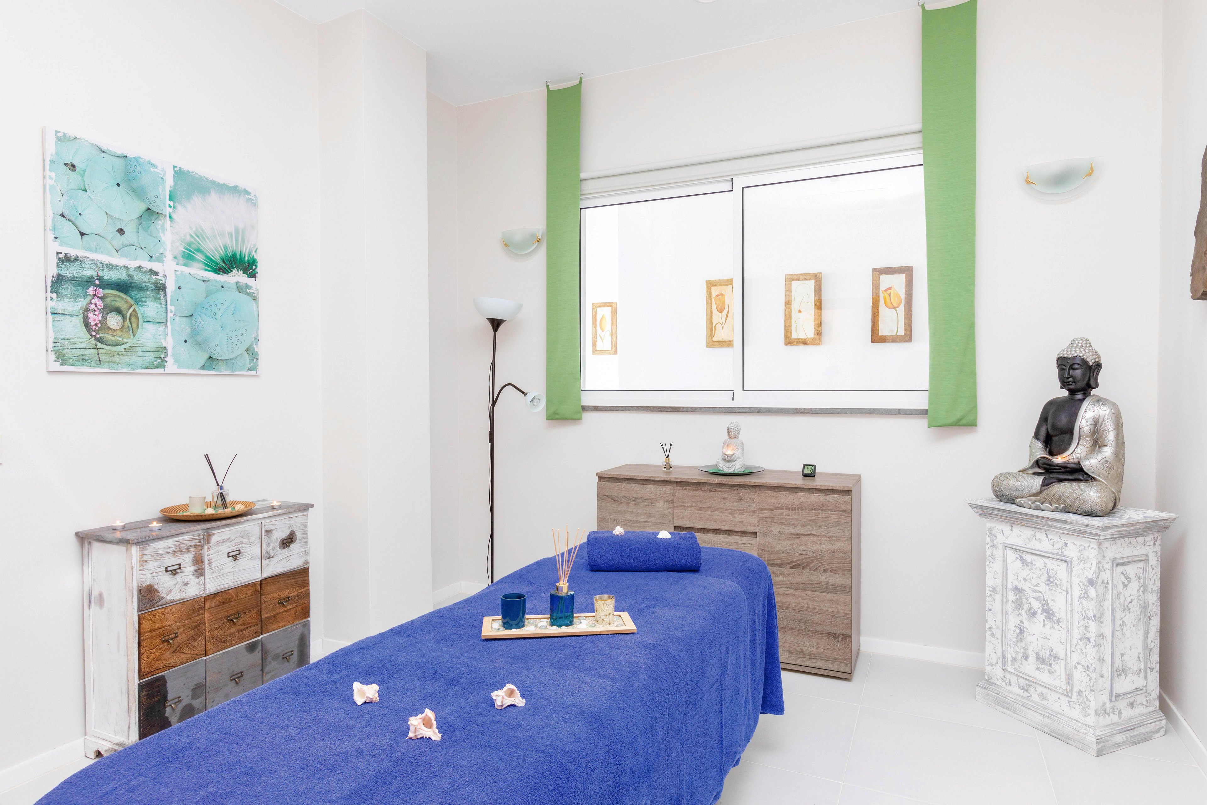 A serene spa treatment room features a blue massage table as the central focus. The setting includes minimalist decor, a Buddha statue, and calming artwork on the walls. Aromatherapy items and candles are arranged on a tray, enhancing the tranquil atmosphere. The room is bright with natural light and soft green accents, creating a peaceful environment for relaxation.