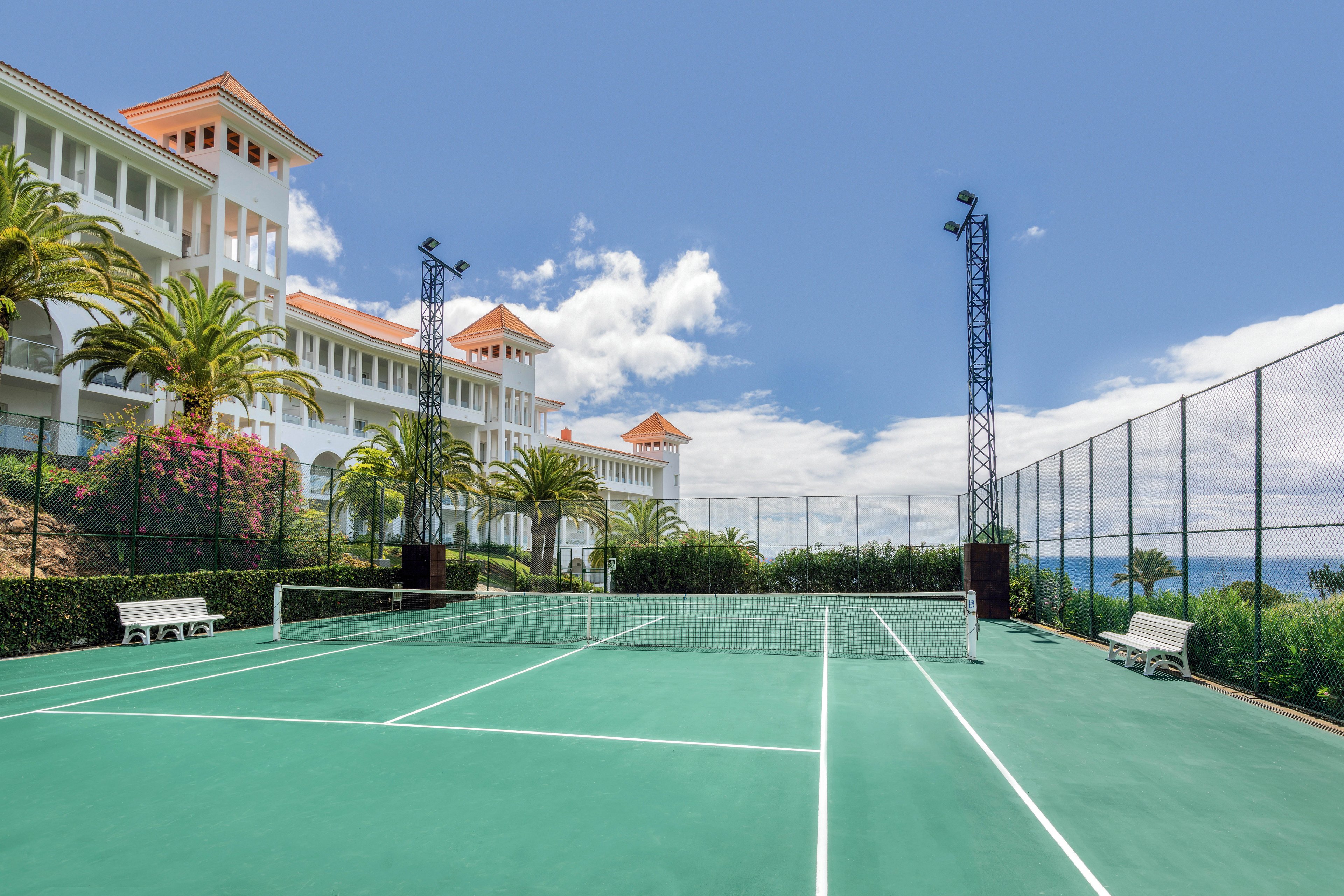 An outdoor tennis court is situated beside a grand resort building with white walls and terracotta roofs. The court is surrounded by lush greenery, palm trees, and flowering bushes, with benches placed along the sides. The scene is brightly lit under a clear blue sky, creating a vibrant and inviting atmosphere.