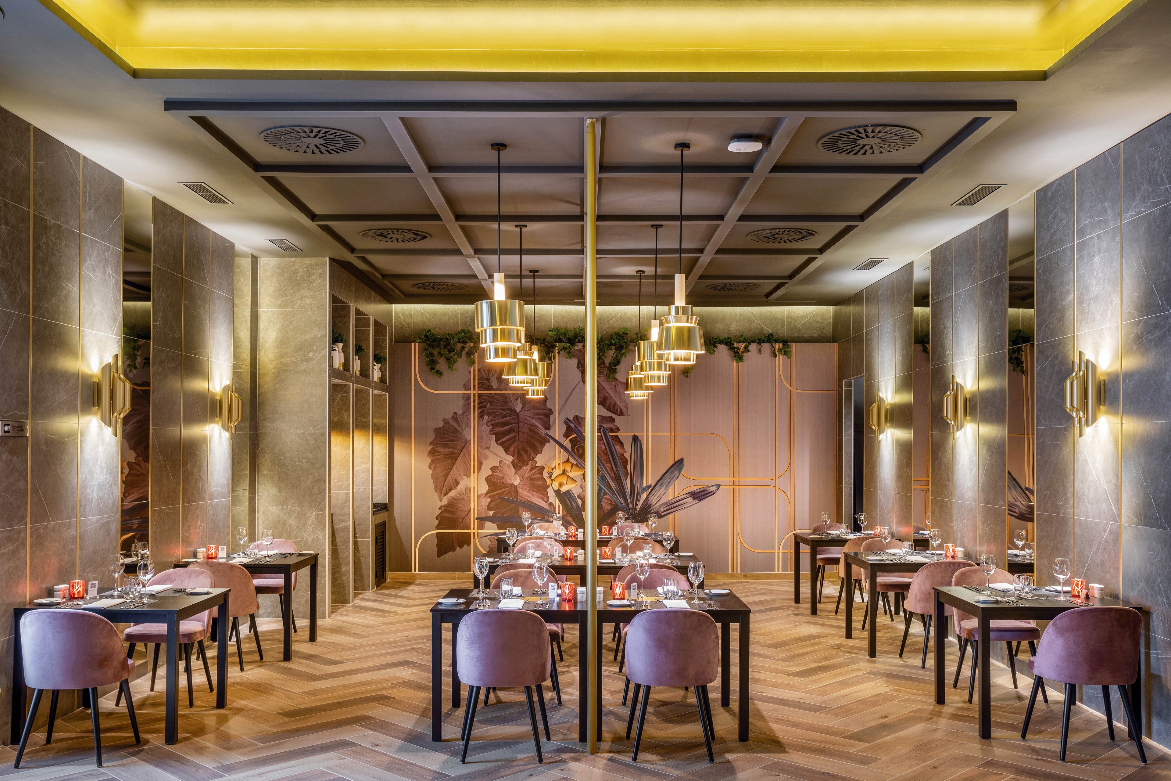 The image shows a contemporary restaurant dining area featuring plush pink chairs and dark wooden tables. The setting is stylish, with geometric ceiling panels, gold pendant lights, and a botanical mural on the back wall. The mood is sophisticated and inviting, with warm lighting and a neutral color palette. No visible text or numeric values are present in the scene.
