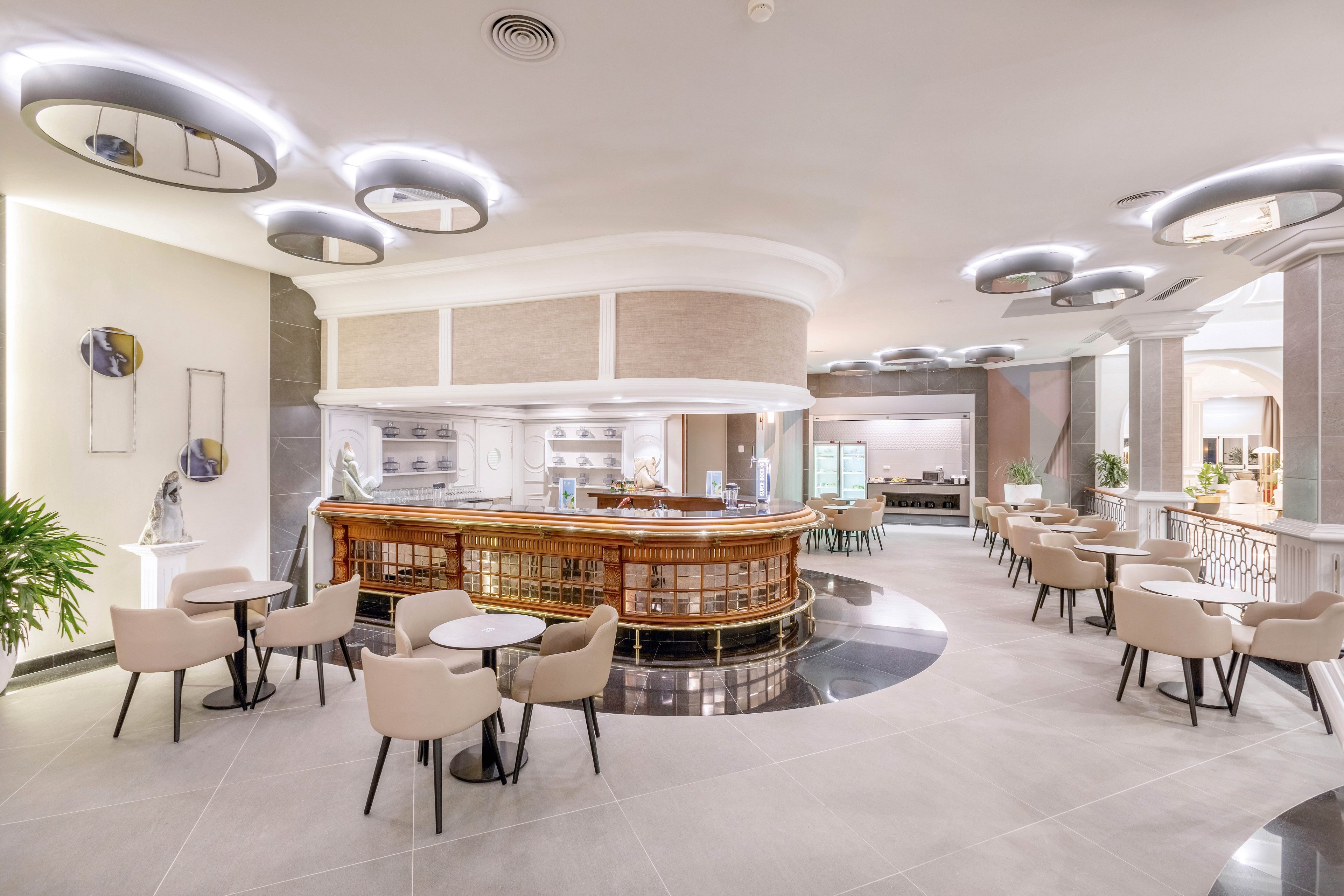 A spacious, contemporary hotel lounge features a central circular bar surrounded by beige chairs and small tables. The setting is bright with neutral tones, modern lighting fixtures, and minimalist decor. The area is empty, emphasizing the clean lines and open layout. Decorative wall art and plants add subtle visual interest to the sophisticated atmosphere.