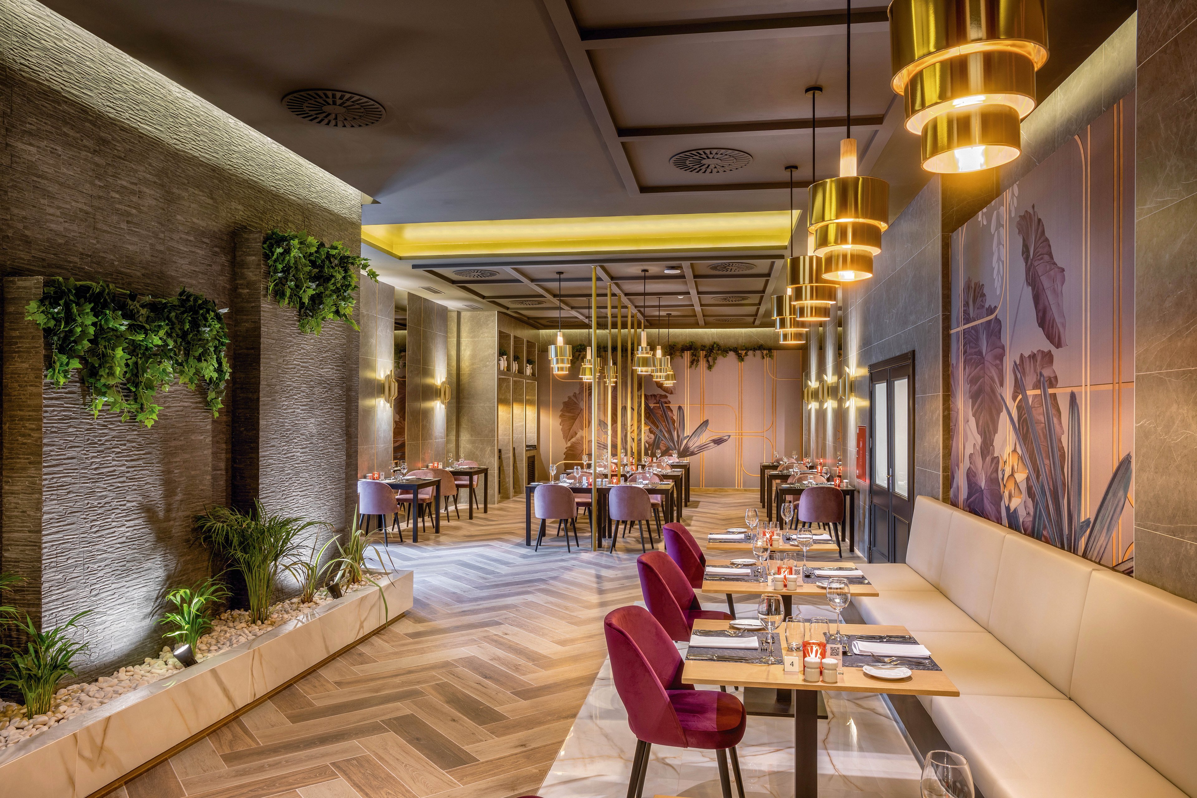 A contemporary restaurant dining area features plush seating, gold pendant lights, and a stylish mix of textures. The setting includes wall art, greenery, and neatly set tables, creating a sophisticated ambiance. Warm lighting and a neutral palette are complemented by purple chairs and wood accents. The space appears ready for guests, emphasizing comfort and modern design.