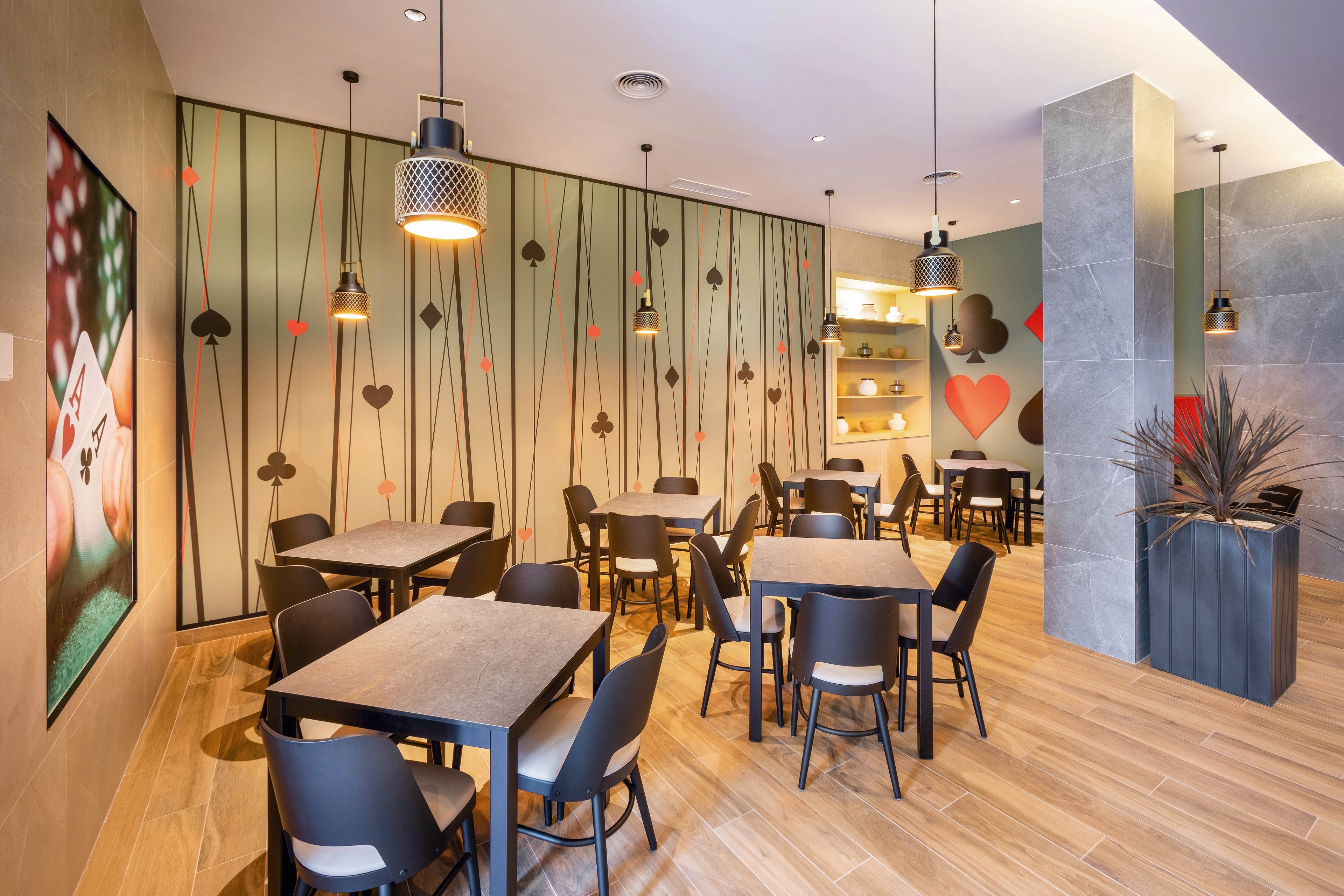 The image shows a contemporary café interior featuring tables and chairs arranged for small groups. The walls are decorated with large playing card motifs, including hearts, spades, clubs, and diamonds, creating a playful and stylish atmosphere. Pendant lights hang from the ceiling, and the space is illuminated with warm tones and natural wood flooring. No visible text or numeric values are present in the image.