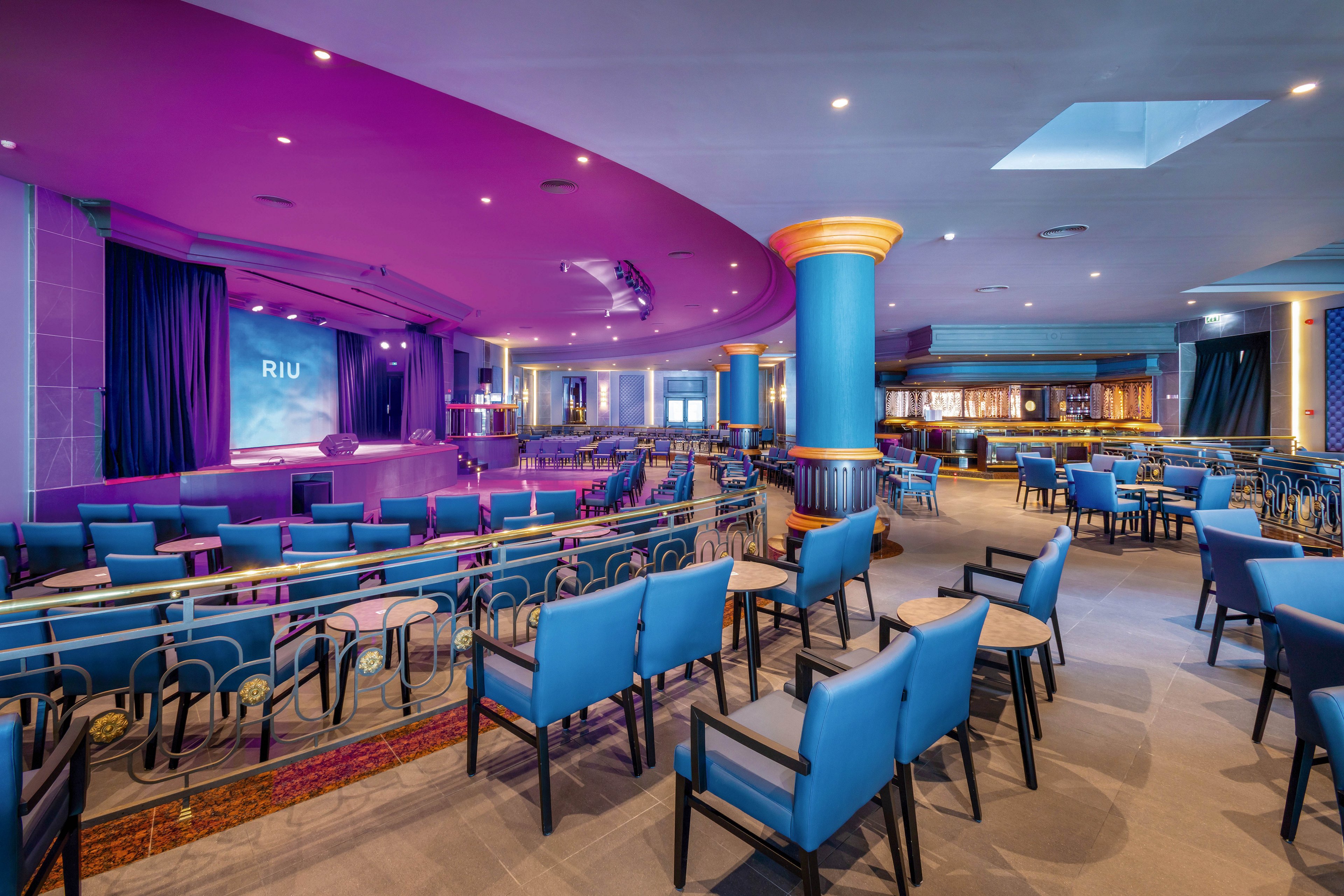 Spacious hotel lounge featuring a stage with the visible text 'RIU' on the screen. The setting includes rows of blue cushioned chairs and tables arranged for an audience, with vibrant purple and blue decor. The area is well-lit, with a bar visible in the background and a large decorative column in the center. The mood is lively and inviting, suitable for entertainment or events.