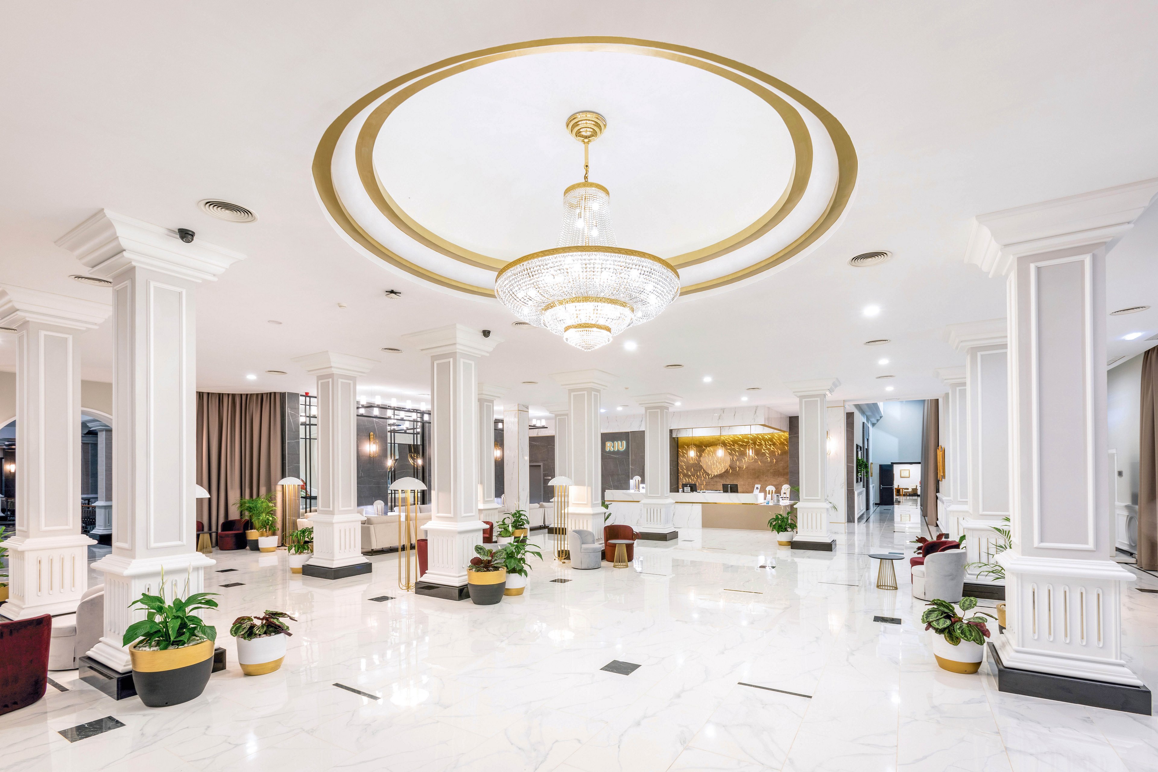 A spacious hotel lobby features a grand chandelier centered in a circular ceiling recess. The area is adorned with white columns, polished marble floors, and modern seating arrangements. Potted plants add greenery throughout the bright, well-lit space. The reception desk is visible in the background, contributing to the welcoming atmosphere.
