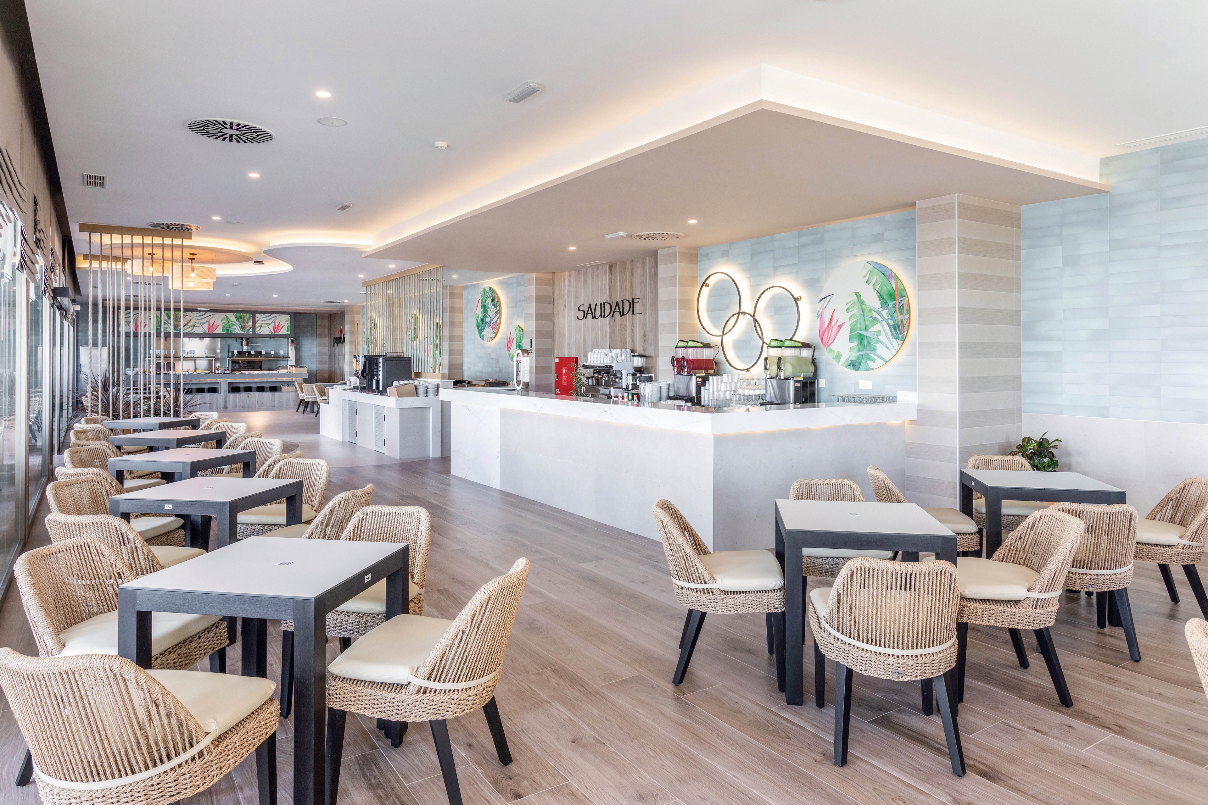 A spacious, contemporary café features light wood flooring, black tables, and woven wicker chairs arranged neatly throughout the room. The counter area is accented by illuminated wall art and a visible 'SALUD!' sign, with coffee machines and beverage dispensers on display. The setting is airy and inviting, with a neutral palette and soft lighting creating a relaxed, welcoming mood.