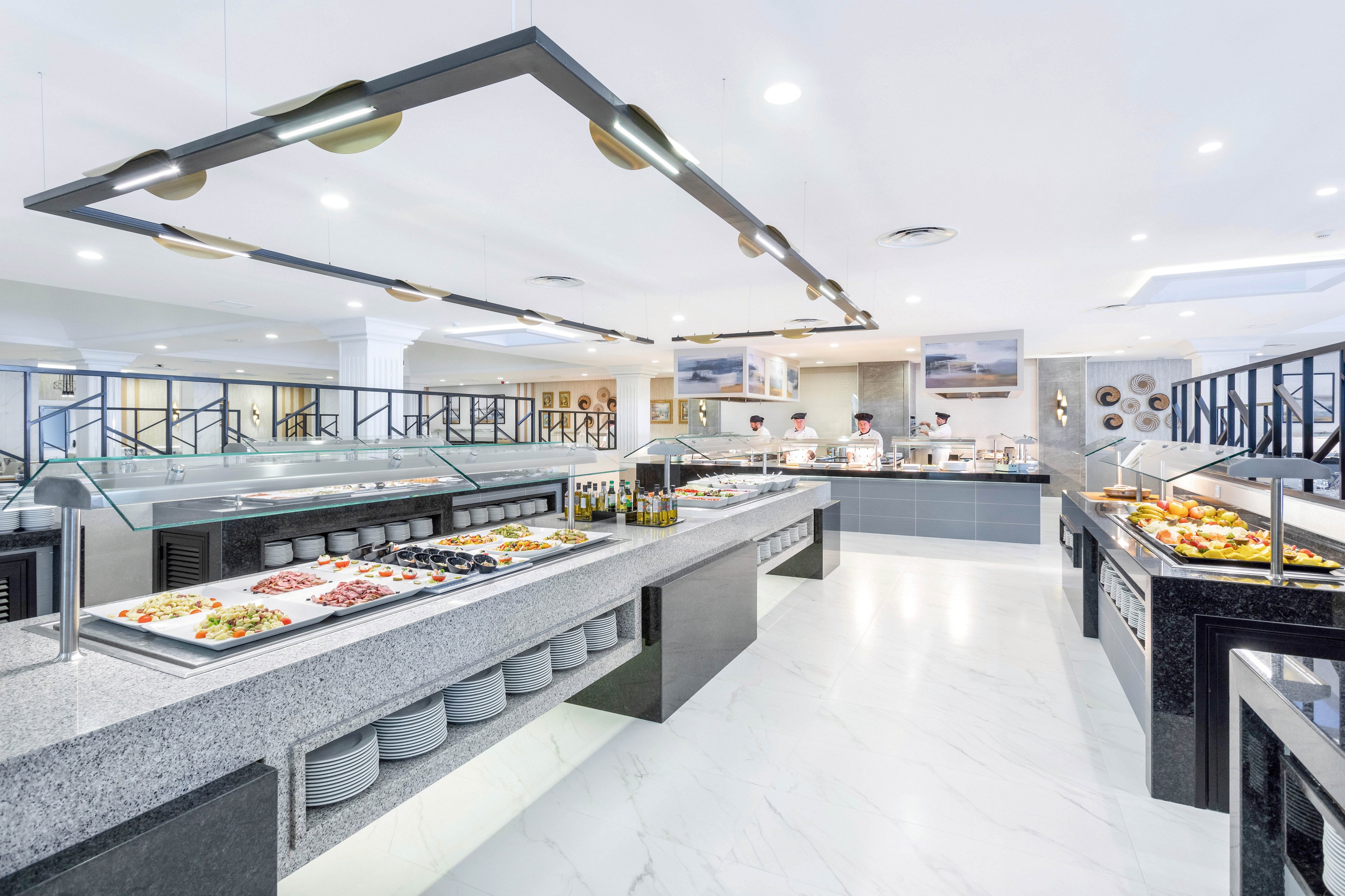 A spacious, contemporary buffet restaurant features long granite counters filled with assorted salads, fruits, and prepared dishes. The setting is bright and clean with white marble floors and minimalist decor. Several chefs are visible in the background preparing food, and the overall mood is inviting and fresh. Plates are neatly stacked under the counters, and the lighting is modern and geometric.