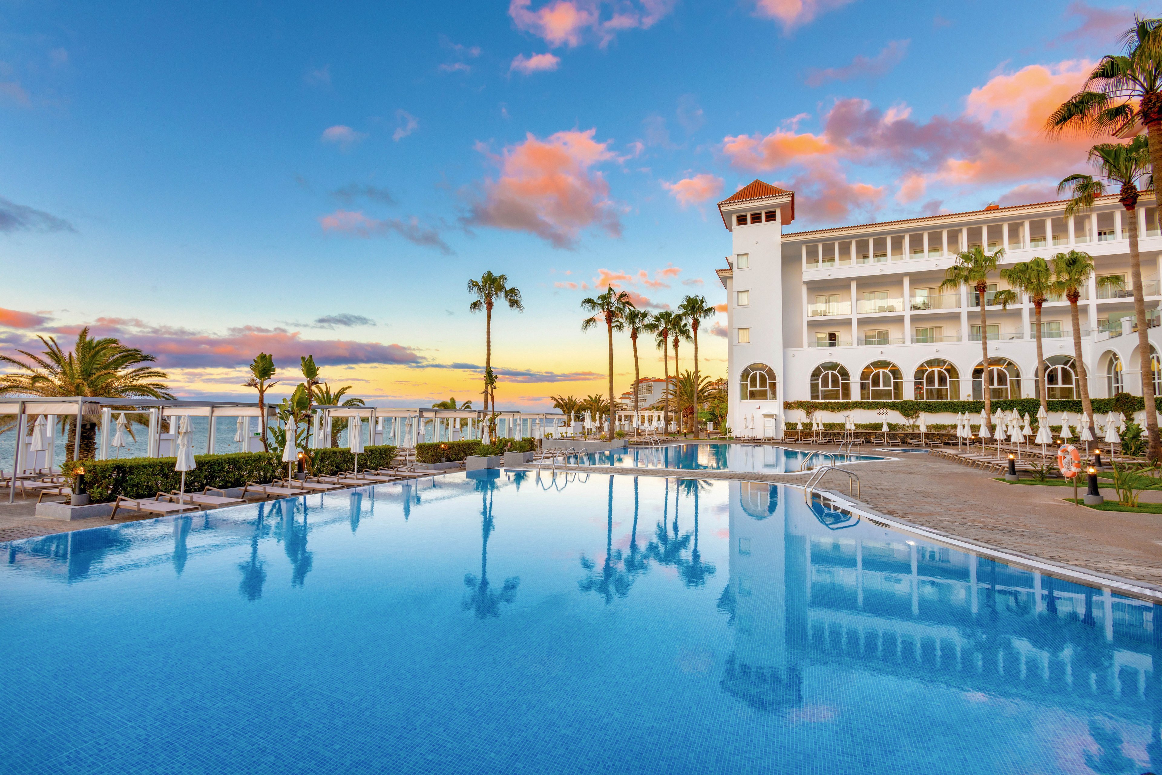 A grand resort hotel stands beside a spacious outdoor swimming pool, reflecting the vibrant colors of a sunset sky. Palm trees and elegant white cabanas line the pool area, creating a tranquil and inviting atmosphere. The building features arched windows and a tower, enhancing the upscale ambiance. The scene is calm, with no visible people, and the palette is dominated by soft blues, pinks, and warm golden tones.
