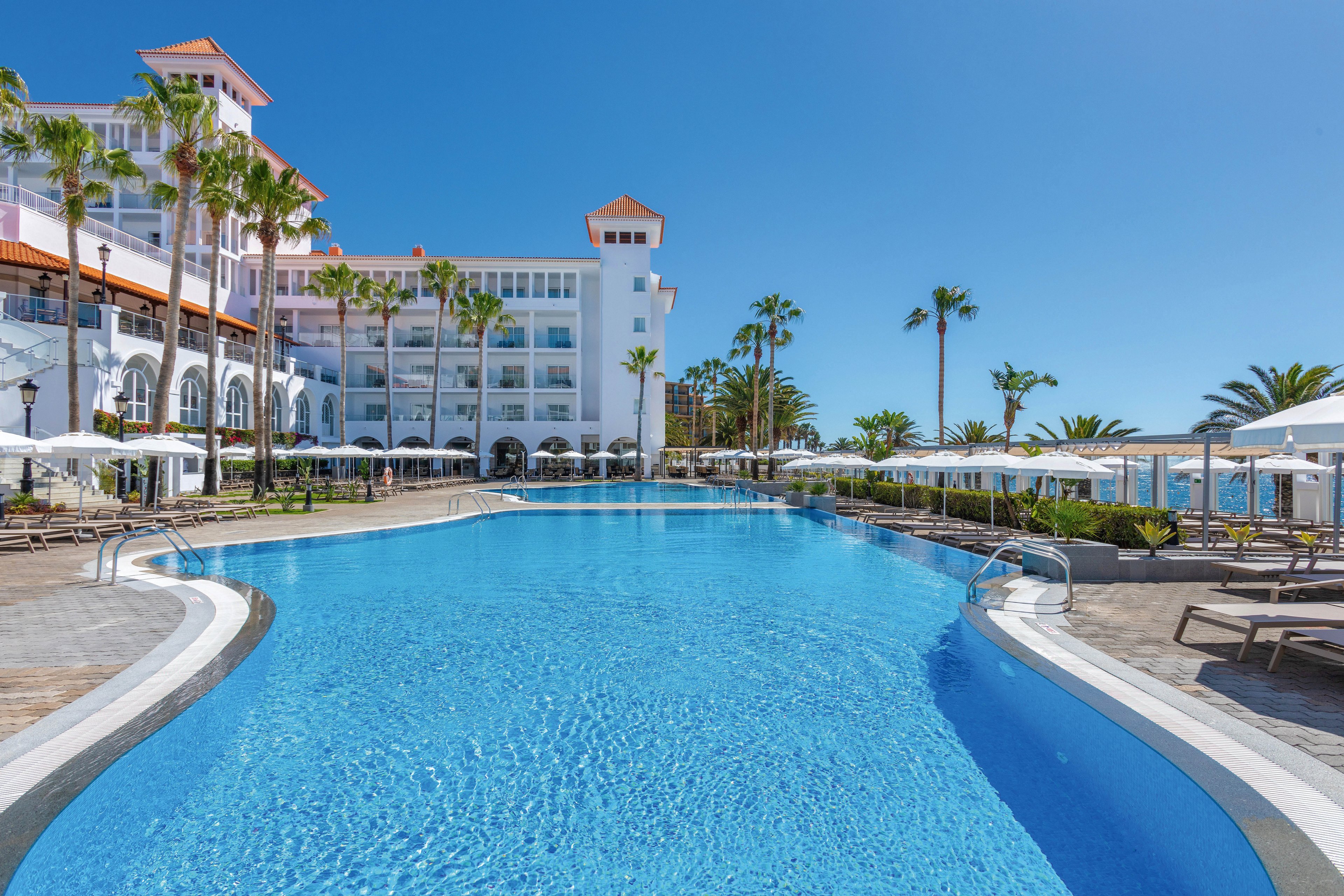 A spacious outdoor swimming pool is the centerpiece of a luxury resort hotel under a clear blue sky. The scene features palm trees, sun loungers, and white umbrellas surrounding the pool, with the hotel building in the background. The setting is bright and inviting, evoking a relaxing vacation atmosphere. No visible text or numbers are present in the image.