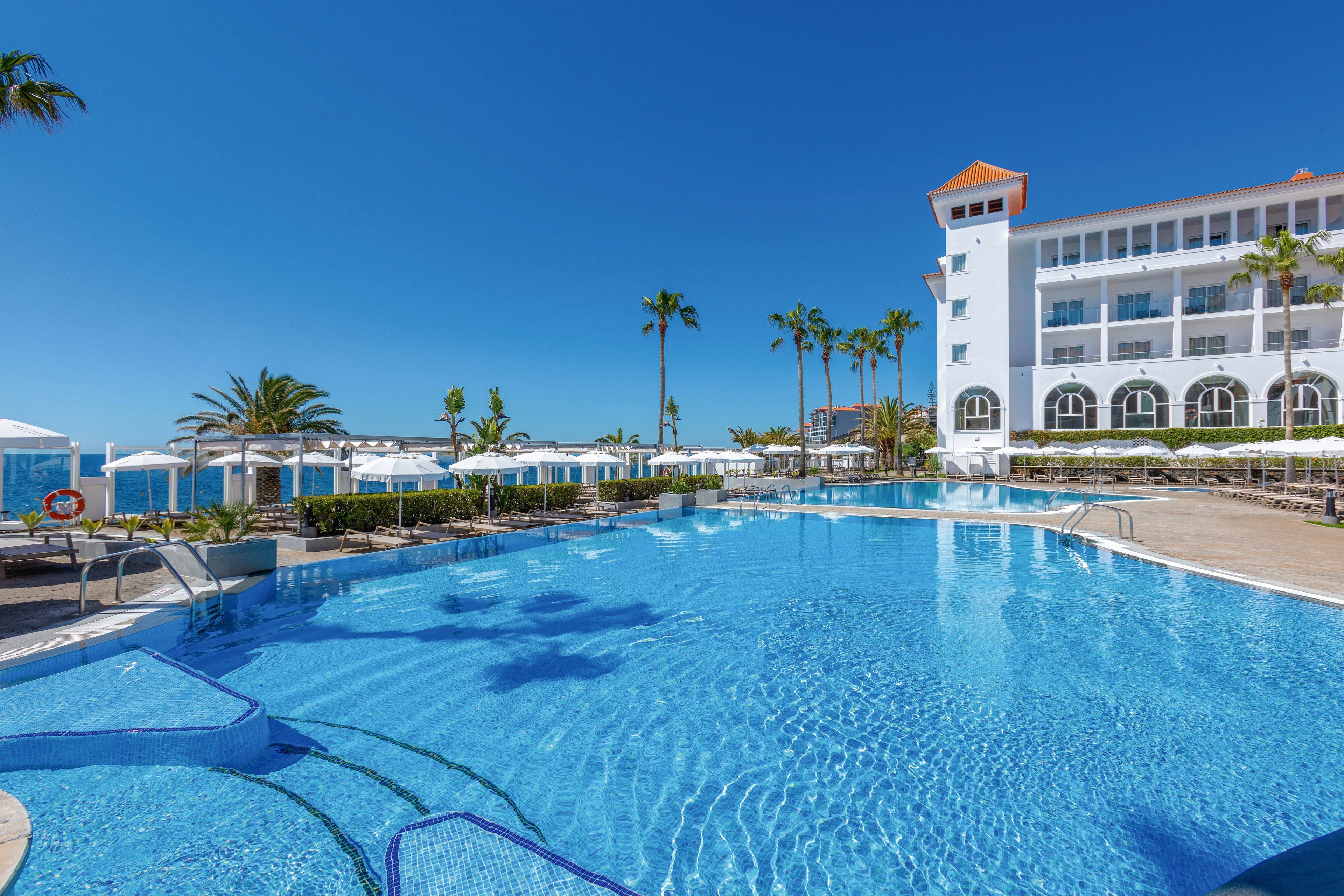A large, inviting swimming pool is the main focus, surrounded by sun loungers and umbrellas at a modern resort. The setting features tall palm trees, clear blue water, and a white multi-story hotel building with arched windows. The atmosphere is bright and tranquil, with a vivid blue sky and no visible people. A life preserver with the number '112' is clearly visible near the poolside.