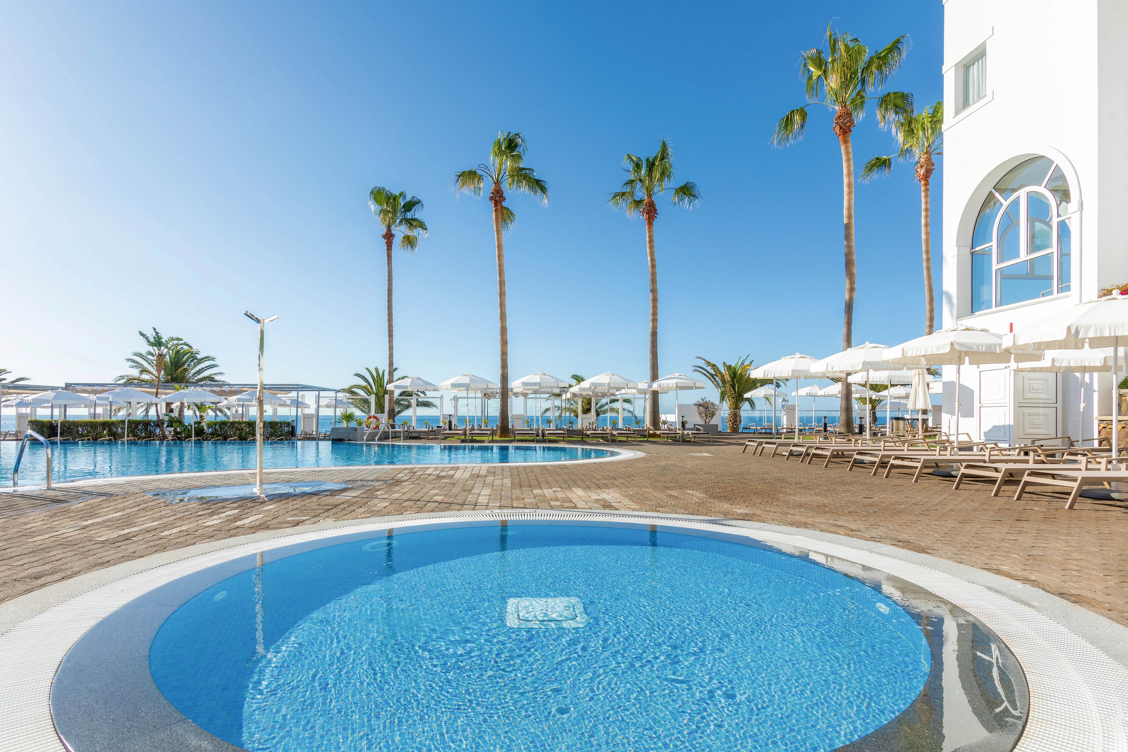 A bright, modern resort pool area features a circular wading pool in the foreground and a larger swimming pool in the background. Tall palm trees line the deck, and numerous sun loungers and white umbrellas are arranged neatly around the pools. The setting is outdoors under a clear blue sky, evoking a relaxed and luxurious vacation atmosphere.