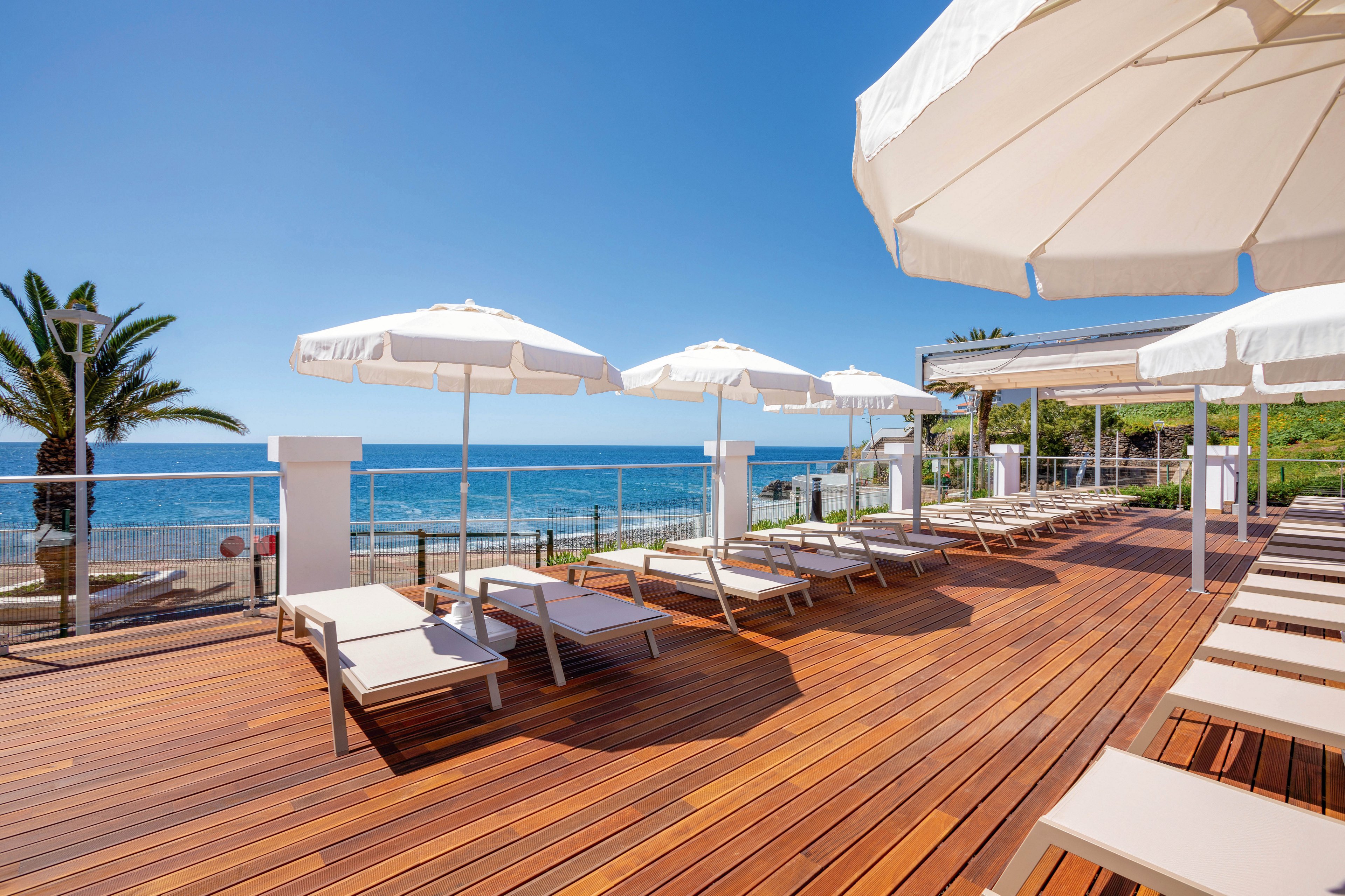 The image shows a spacious wooden deck overlooking the ocean, featuring several lounge chairs and large white umbrellas. The setting is bright and sunny, with clear blue skies and palm trees visible in the background. The mood is relaxed and inviting, ideal for leisure and vacation. No visible text or numeric values are present in the image.