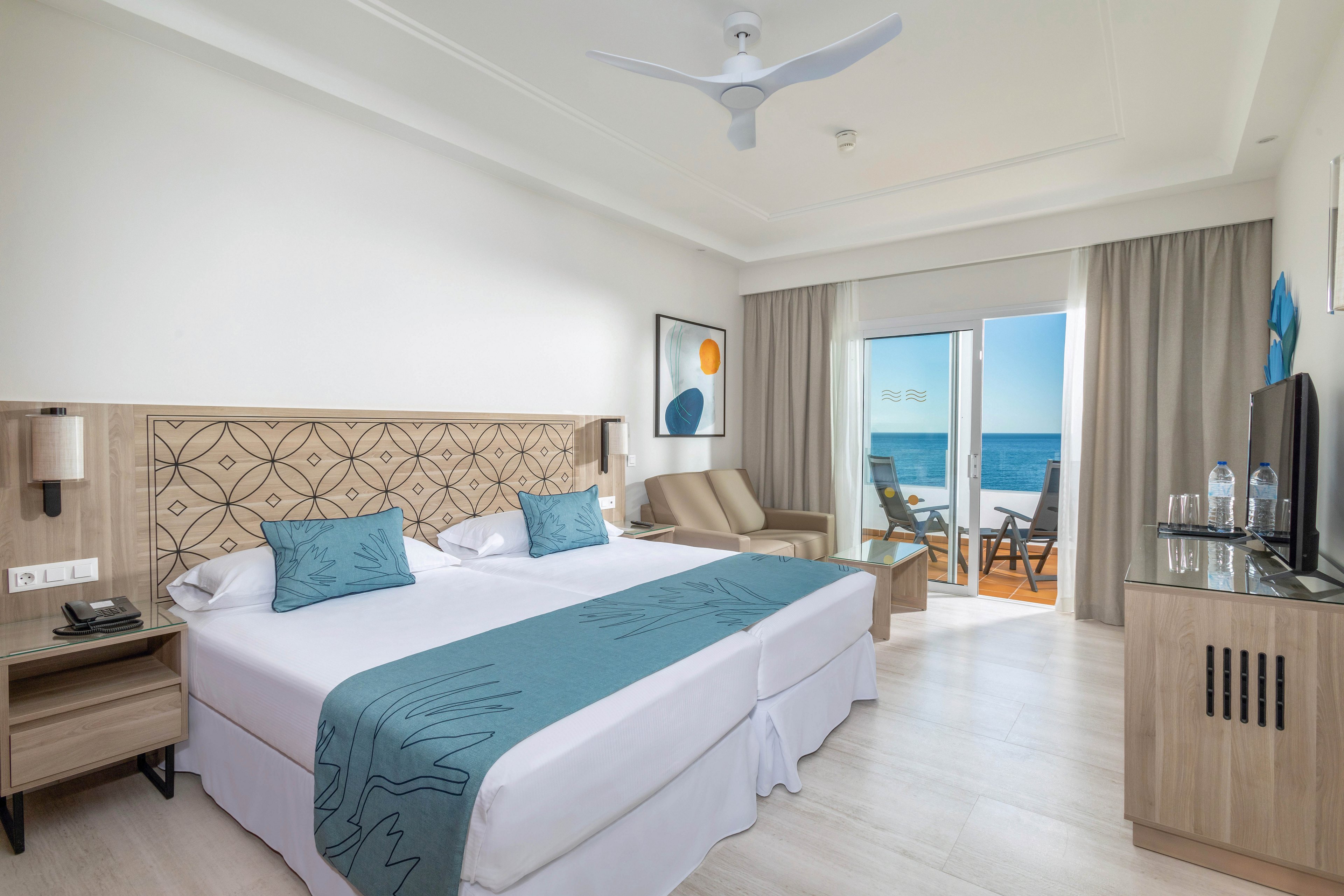 A contemporary hotel room features two twin beds with teal accents, a stylish headboard, and a seating area. The space is bright and airy, with large glass doors opening to a balcony overlooking the ocean. Minimalist decor, neutral tones, and natural light create a relaxing atmosphere. Bottled water and a flat-screen TV are visible on the cabinet.