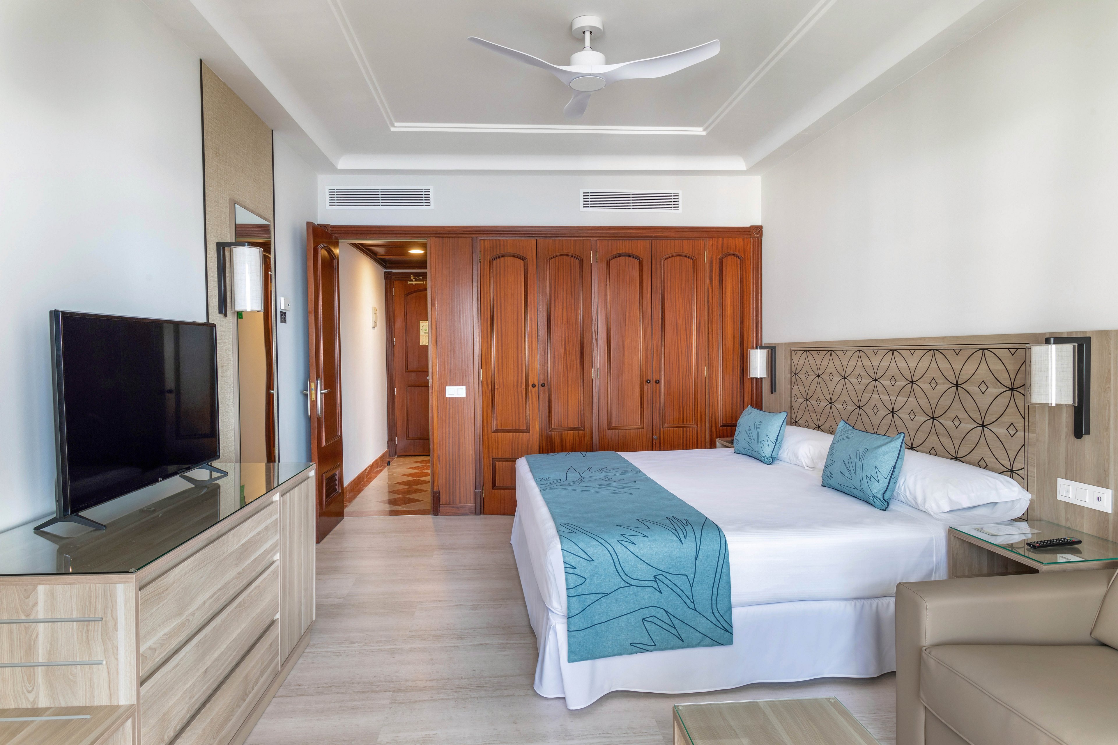 A spacious hotel room features a large bed with blue decorative pillows and a matching runner. The setting includes wooden wardrobes, a flat-screen television, and neutral-toned furniture, creating a clean and inviting atmosphere. The room is well-lit with natural light and has a ceiling fan for comfort. No visible numeric values or identifiable brands are present.