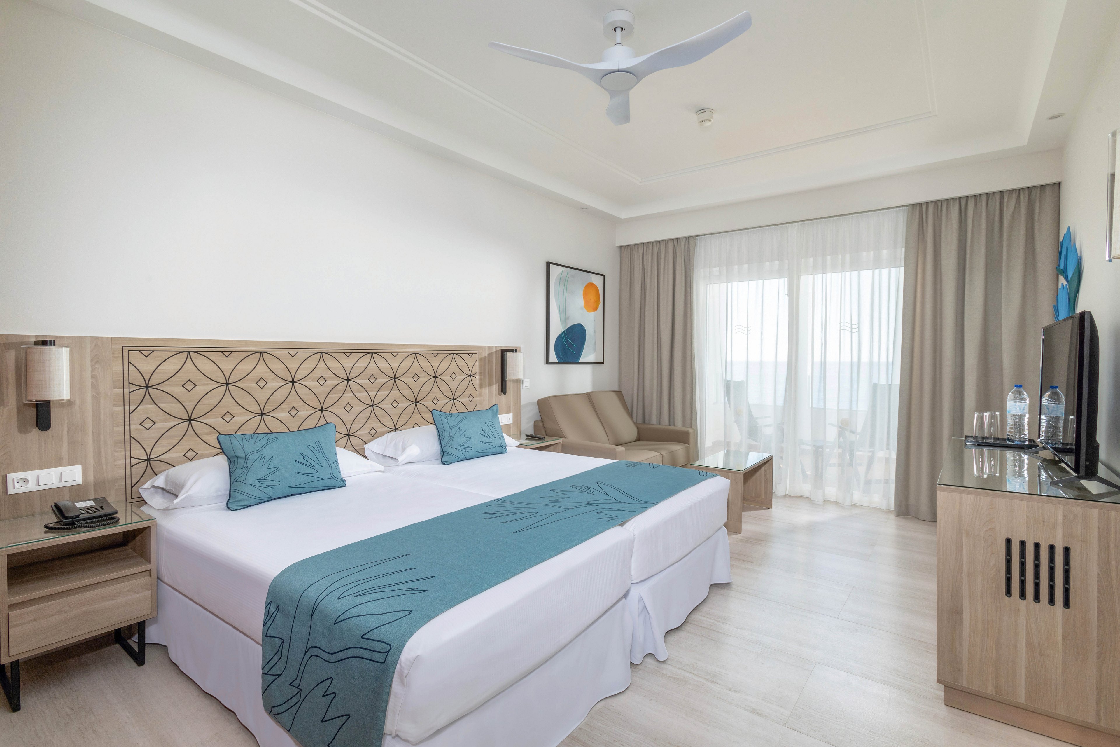 A contemporary hotel room features two twin beds with teal accents and geometric headboard. The space includes a sitting area, flat-screen TV, bottled water, and a balcony with sheer curtains letting in natural light. Minimalist decor and neutral palette create a calm, inviting atmosphere. No visible numeric values or identifiable brands are present.