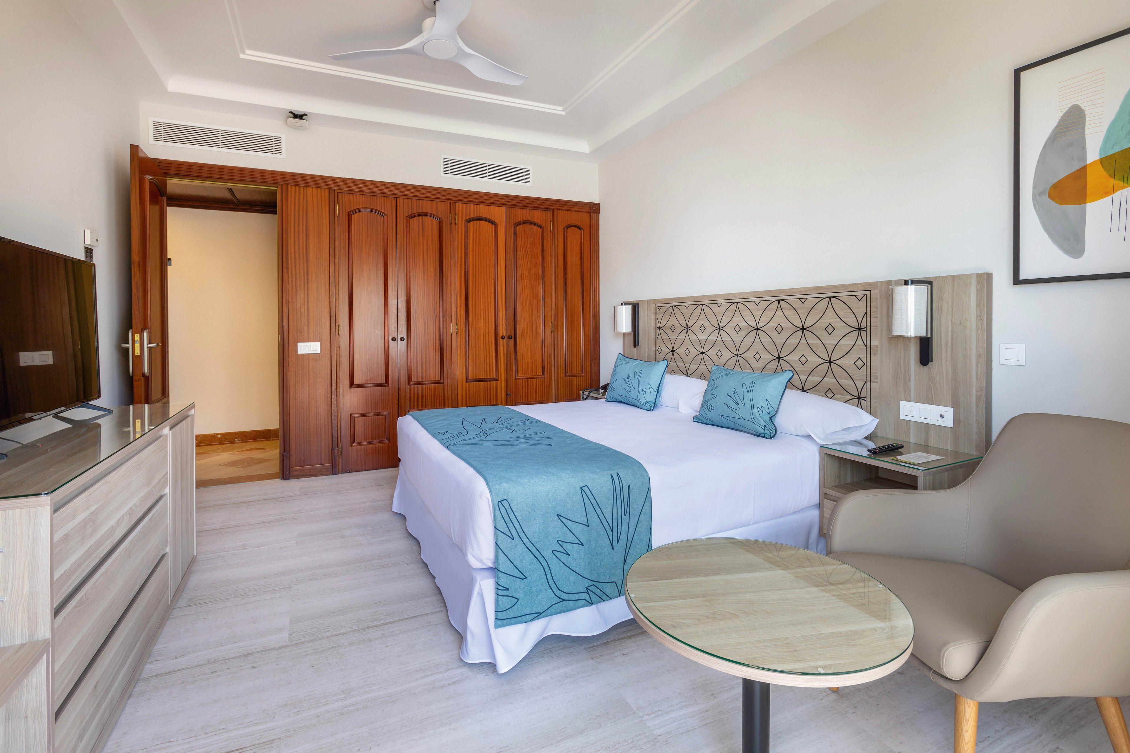 A contemporary hotel room features a double bed with blue accent pillows and a matching bed runner. The space includes a flat-screen television, wooden wardrobe, and a comfortable armchair beside a round table. The room is well-lit with neutral tones, geometric wall art, and clean lines, creating a calm and inviting atmosphere.