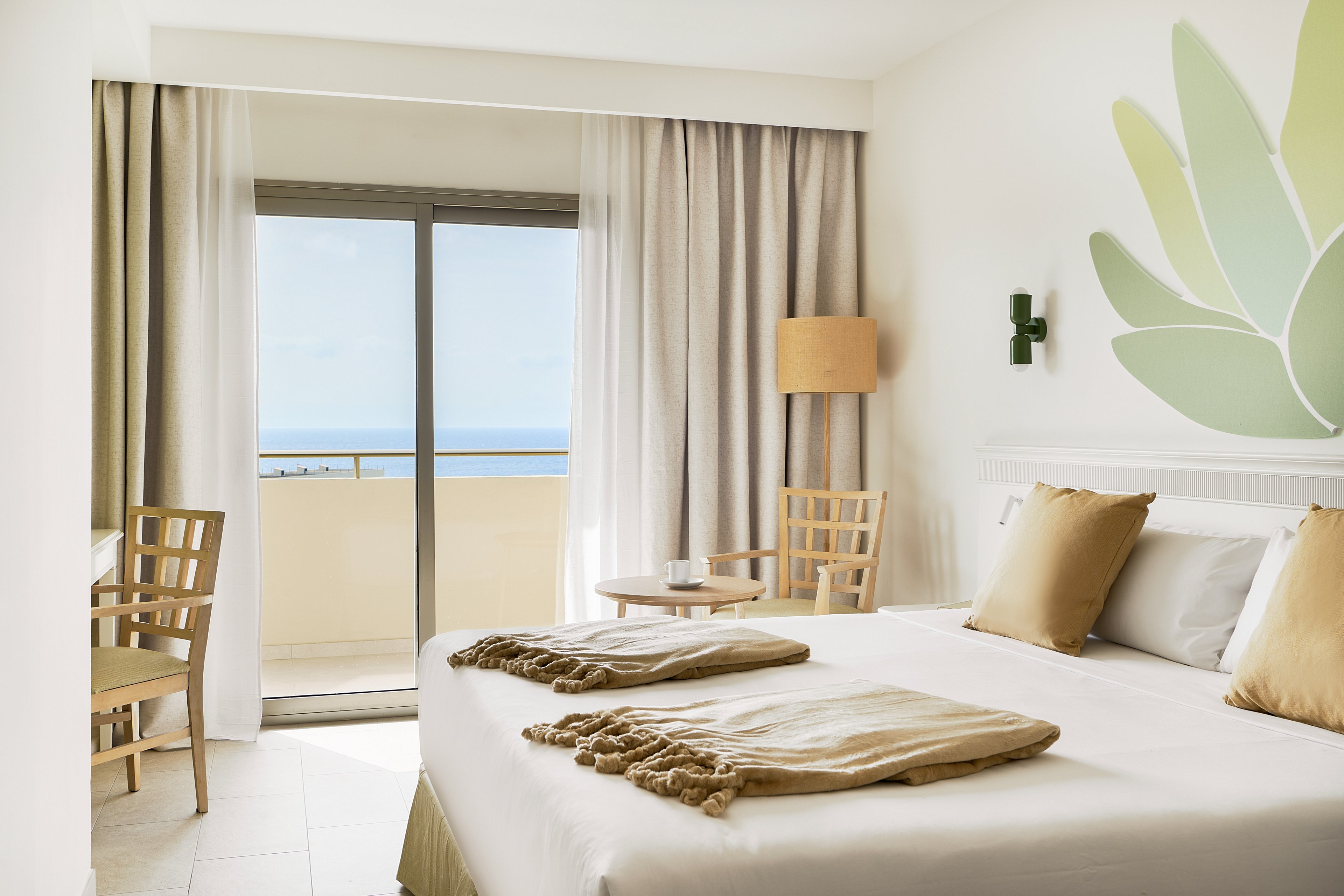 A modern hotel room featuring a neatly made bed with beige pillows and throws. The room is decorated in a minimalist style with light wooden furniture and a green leaf wall design. A sliding glass door opens to a balcony with a view of the ocean under a clear sky. Natural light fills the space, creating a serene and inviting atmosphere.