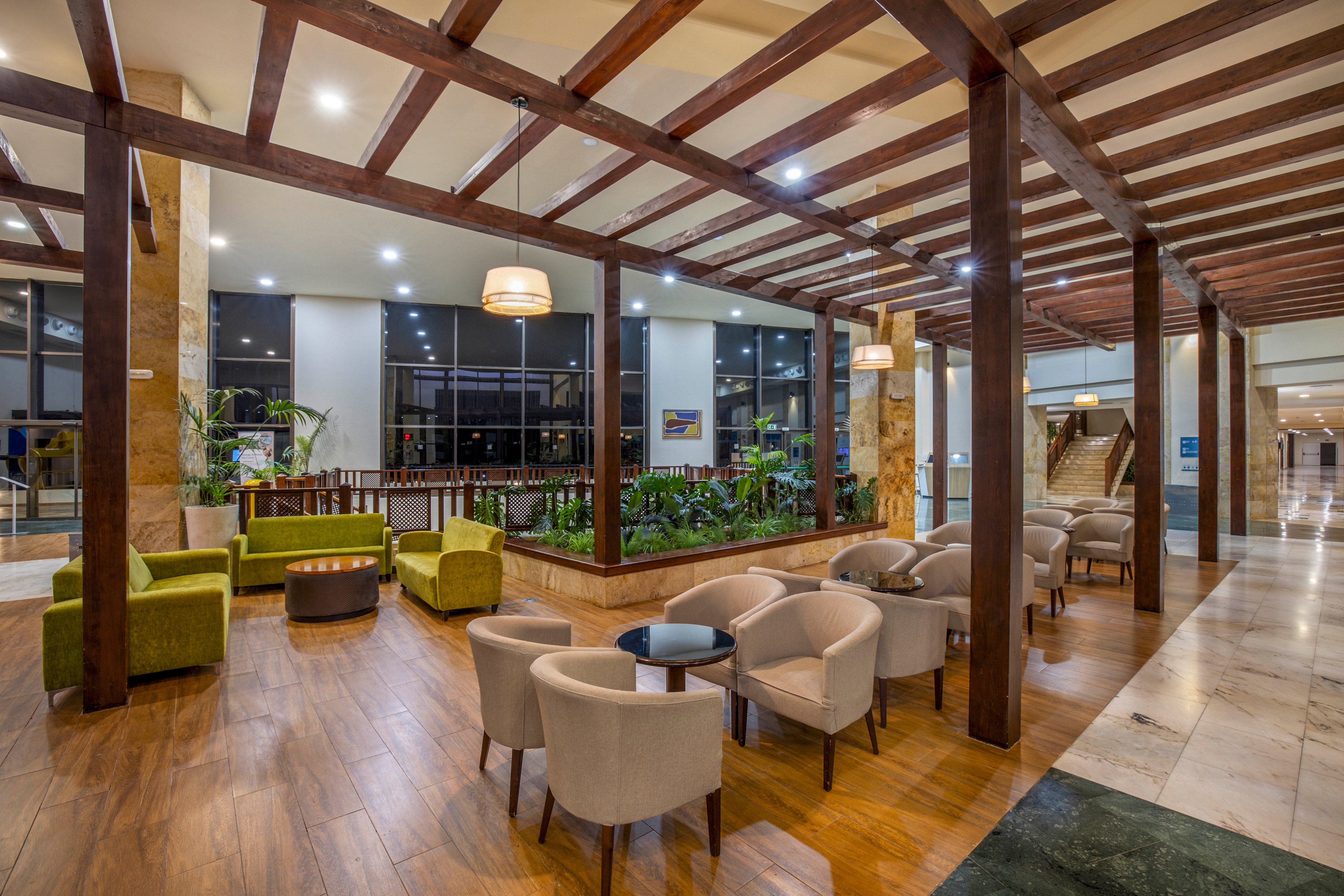A contemporary hotel lobby lounge features multiple seating areas with armchairs and small round tables. The space is defined by a wooden pergola-style ceiling and warm wood flooring. Large windows line the back wall, reflecting interior lights and nighttime views outside. Green upholstered chairs and indoor plants add color and a natural touch to the neutral-toned interior.