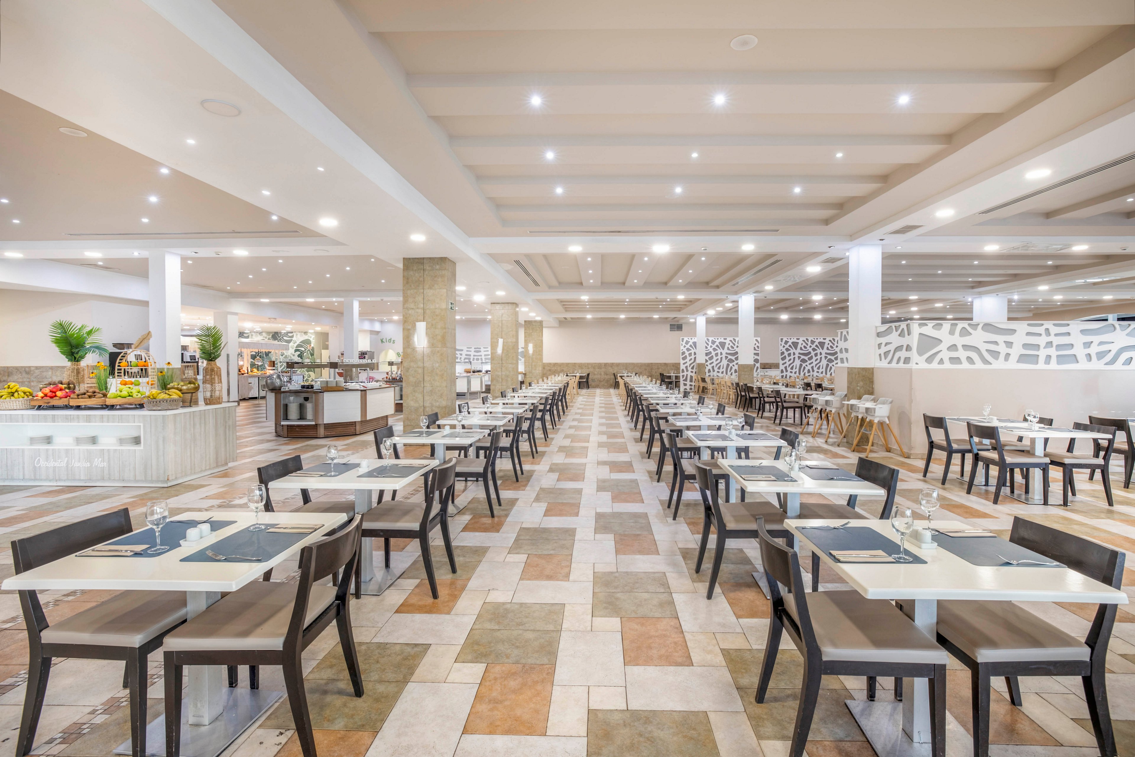 A spacious modern restaurant dining hall features long rows of neatly arranged tables and chairs. The room is brightly lit with recessed ceiling lights and a neutral color palette of beige, white, and light brown. A buffet area with various food displays is visible on the left side of the image. The overall scene is clean, orderly, and ready to welcome guests, though no people are present.