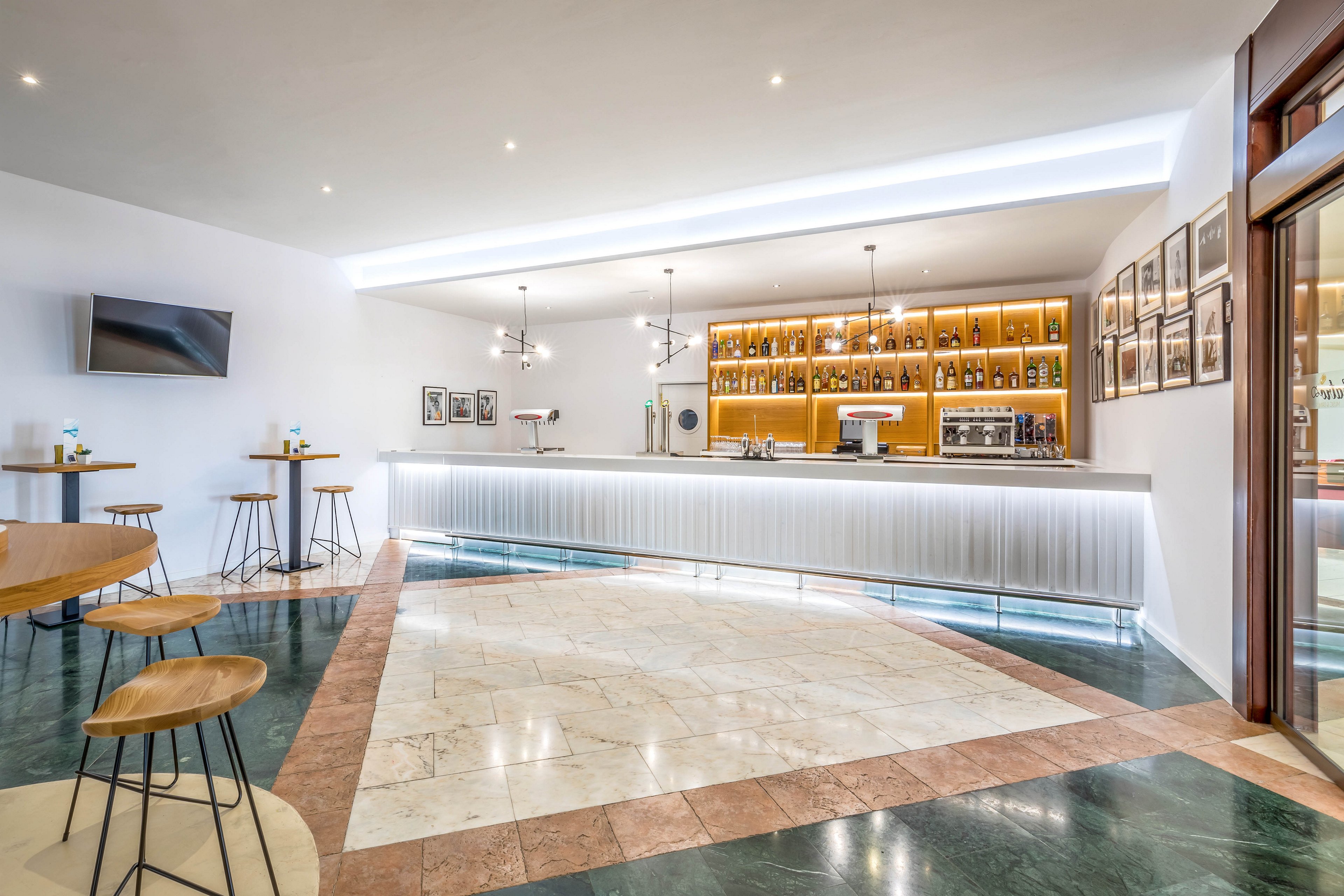 A sleek, modern bar interior features a long white counter with a backlit shelving unit displaying bottles and glassware. High wooden stools and small round tables are arranged around the polished marble and stone floor. Framed photographs line the wall near the bar, while a flat-screen television is mounted on the opposite wall. Recessed ceiling lights and contemporary pendant fixtures create a bright, welcoming atmosphere in the empty space.