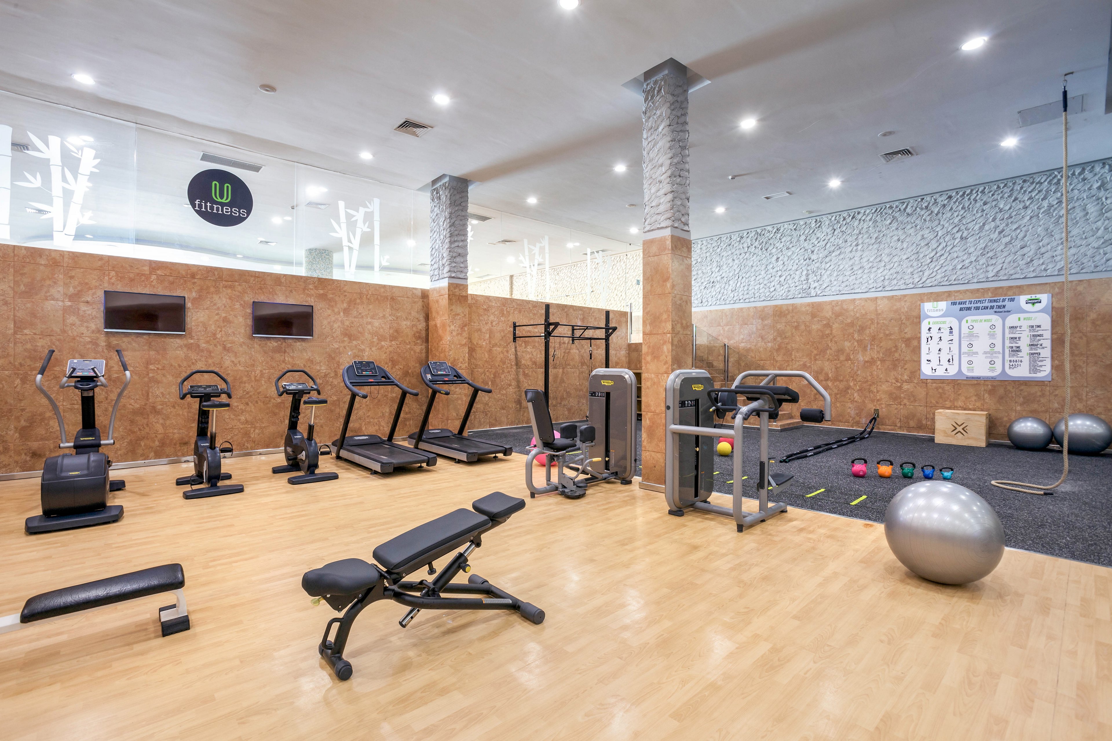 A spacious, modern gym interior features various cardio machines and strength training equipment neatly arranged on a light wooden floor. Treadmills, stationary bikes, and ellipticals line one side, while benches, weight machines, and free weights occupy the center. Exercise balls, kettlebells, and a rope training area are visible near a wall with instructional posters. Bright overhead lighting and neutral brown and white walls create a clean, professional fitness environment.