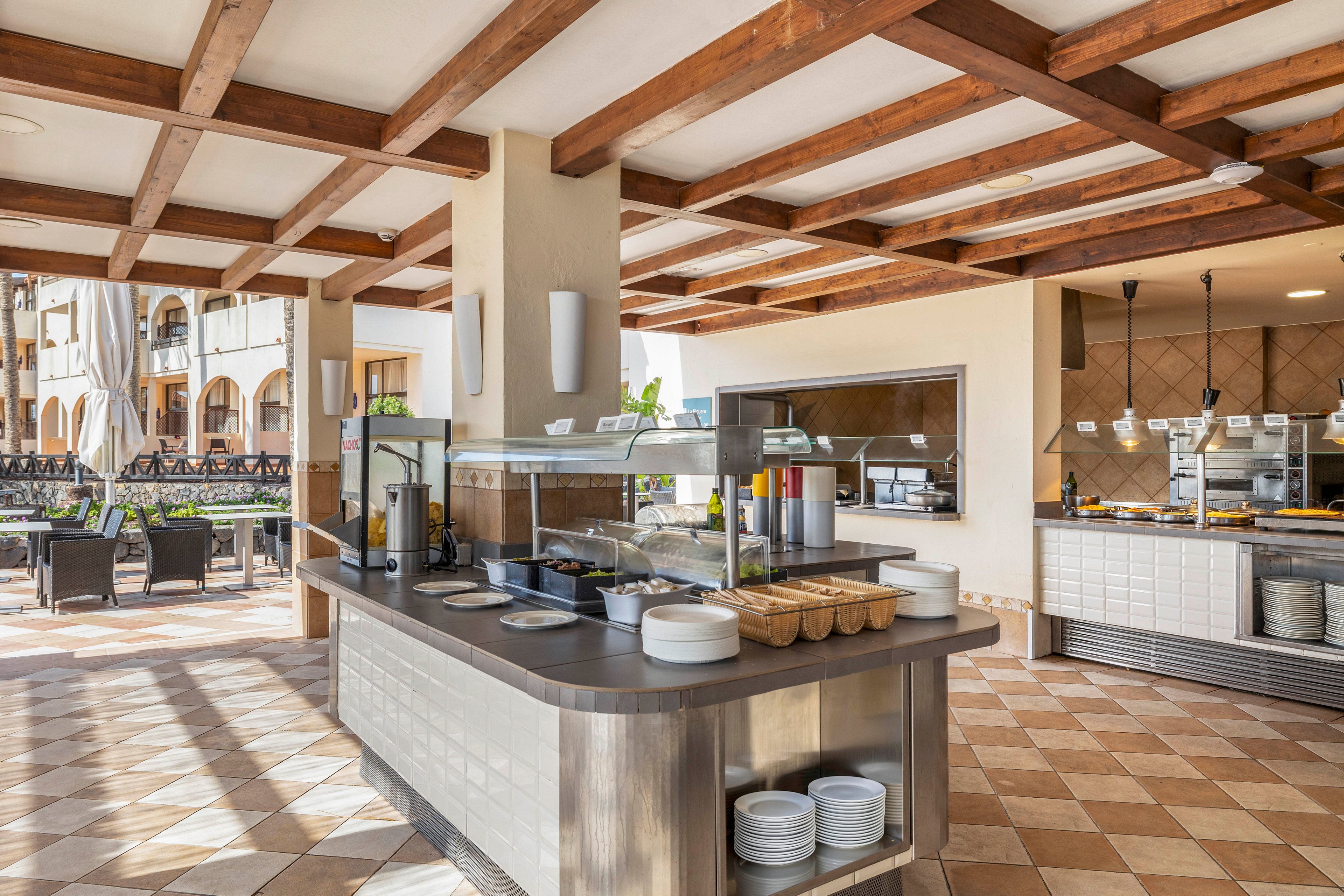 An open-air hotel buffet station is set up under a wooden-beam ceiling. The serving counter displays covered dishes, bowls, and stacked plates ready for guests. In the background, additional food stations and stainless steel equipment are visible. Outdoor dining tables and chairs extend into a sunny courtyard with tiled floors and arched building facades. The scene suggests a relaxed resort atmosphere prepared for meal service.
