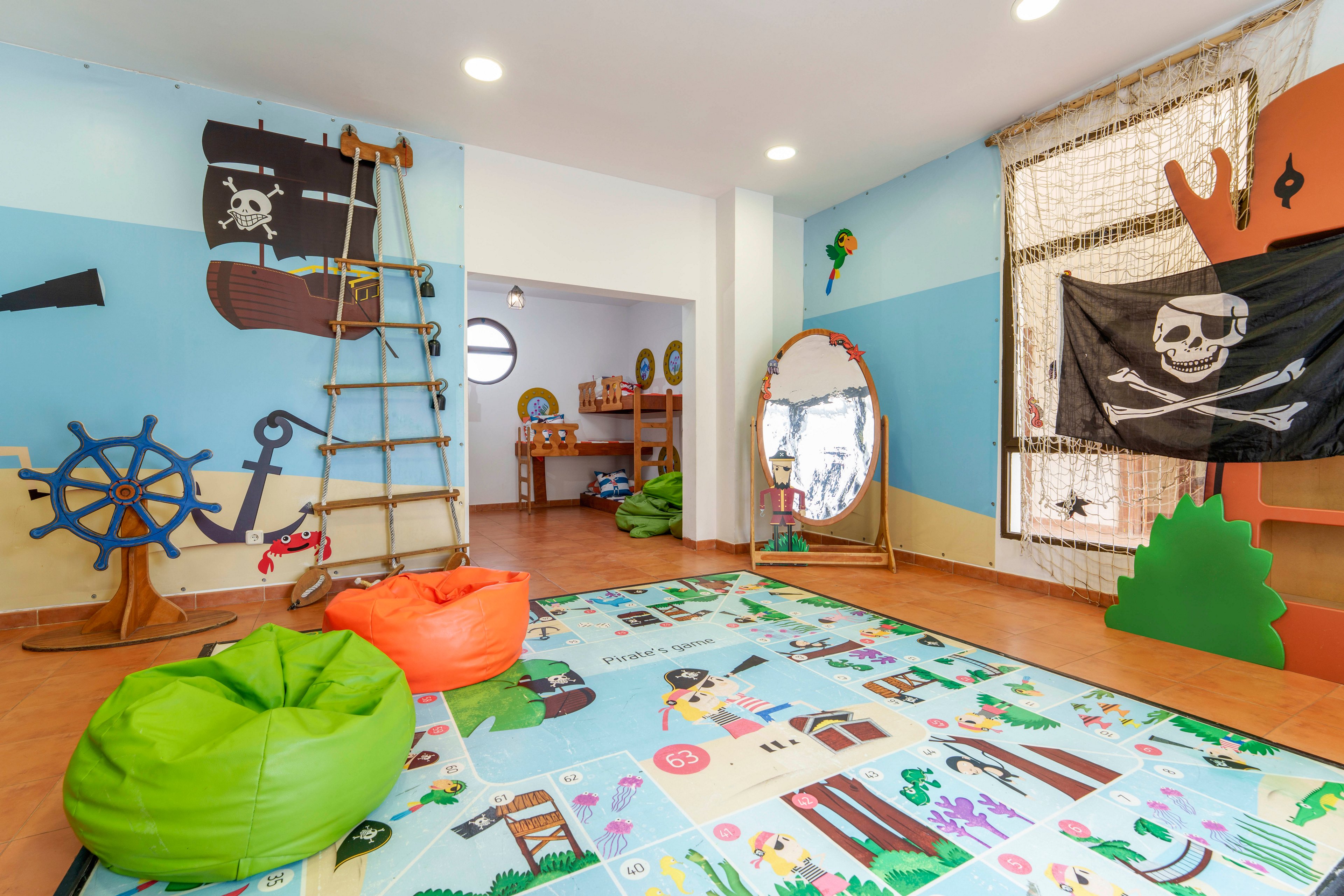 A bright children’s playroom is decorated with a playful pirate theme. The walls feature painted pirate ships, a ship wheel, and nautical elements, while a black pirate flag hangs near a window. Colorful bean bags and a large illustrated play mat with cartoon scenes cover the floor, creating a cozy area for kids to sit and play. In the background, a wooden ladder, a round mirror, and a small table with toys add to the imaginative environment.