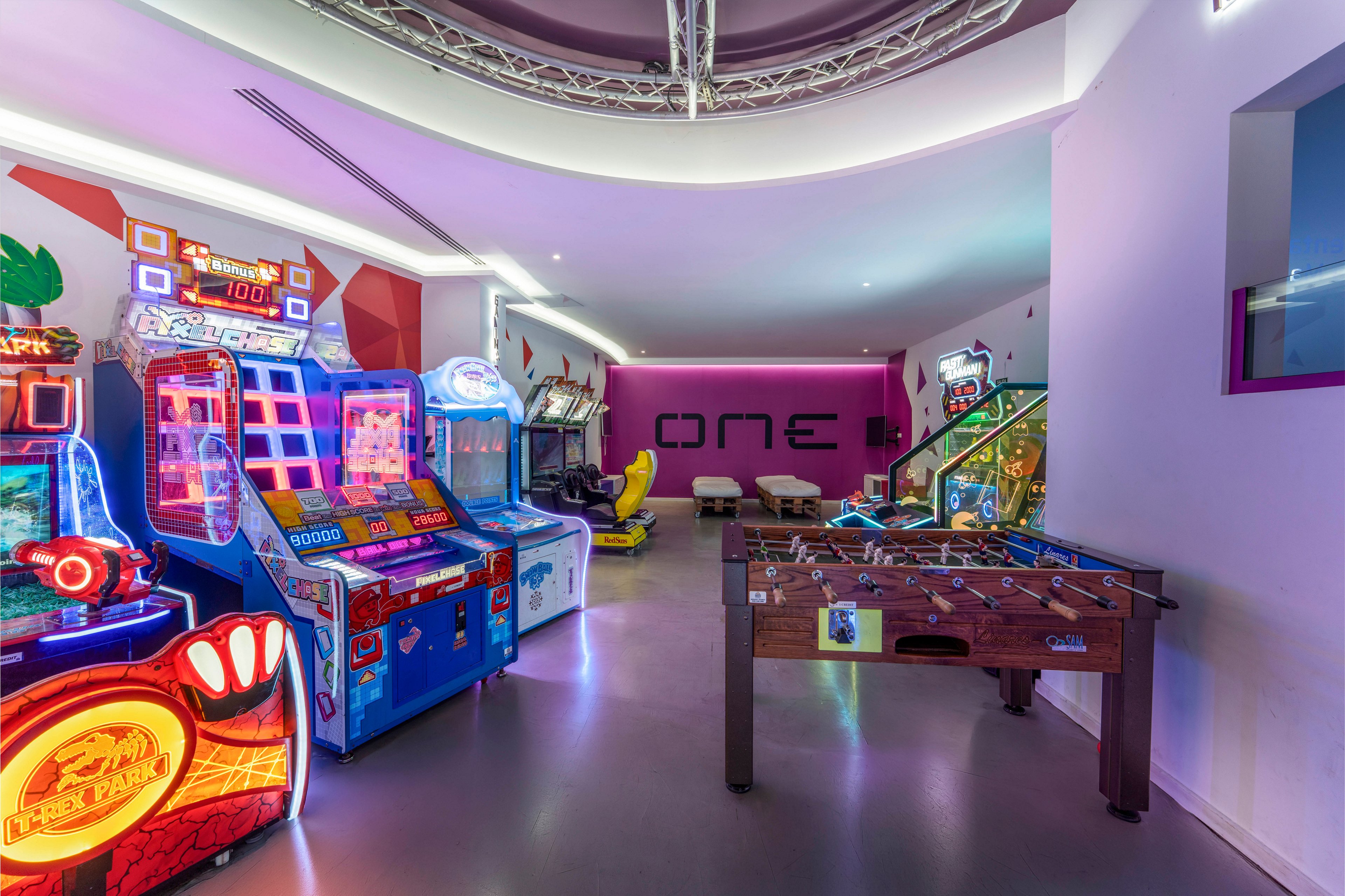 A contemporary arcade room features several brightly lit arcade machines along one wall. In the foreground, a wooden foosball table stands on a polished floor. Neon and LED lights illuminate the space, with a glowing sign reading "ONE" on the back wall. The room has a playful, futuristic atmosphere with colorful reflections and gaming equipment.