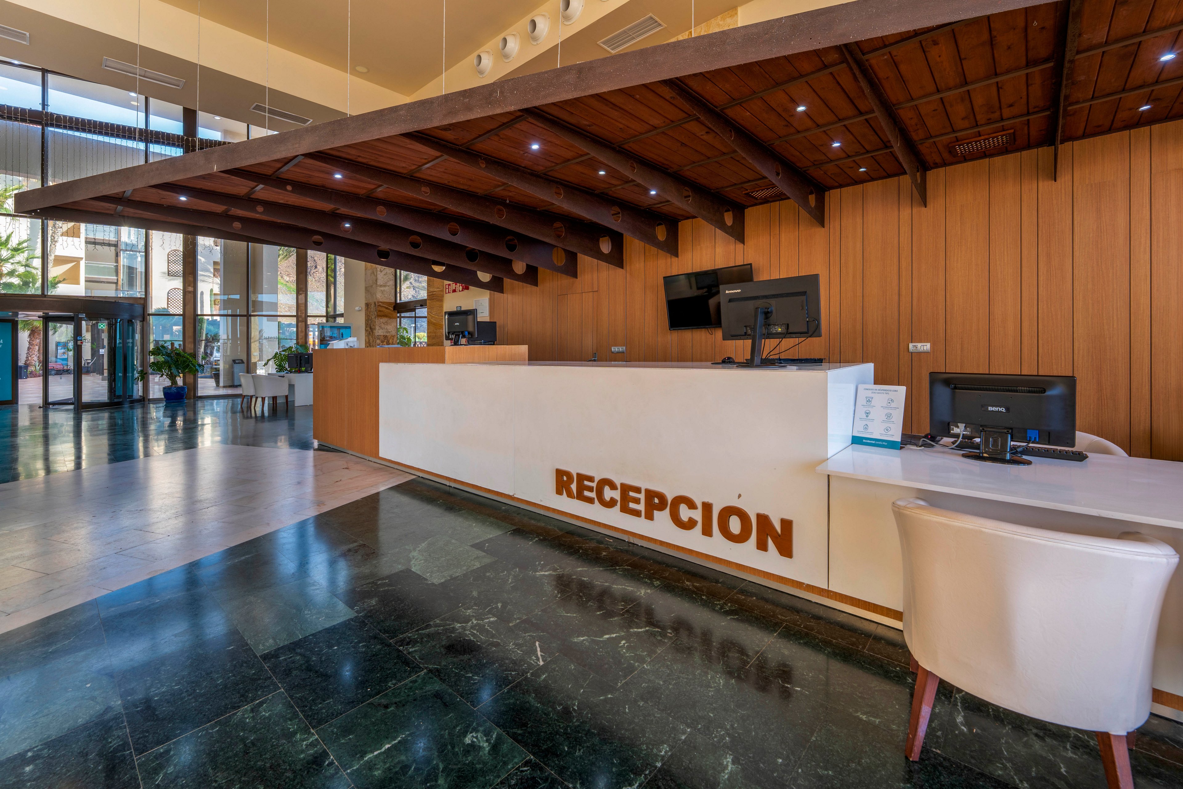 Spacious hotel lobby featuring a large wooden reception desk labeled "RECEPCION". The area includes multiple computer monitors, a telephone, and a white guest chair at the front counter. High ceilings, glass entrance doors, and polished dark flooring create an open, contemporary atmosphere. Natural light and warm wood tones contribute to a welcoming, professional setting.