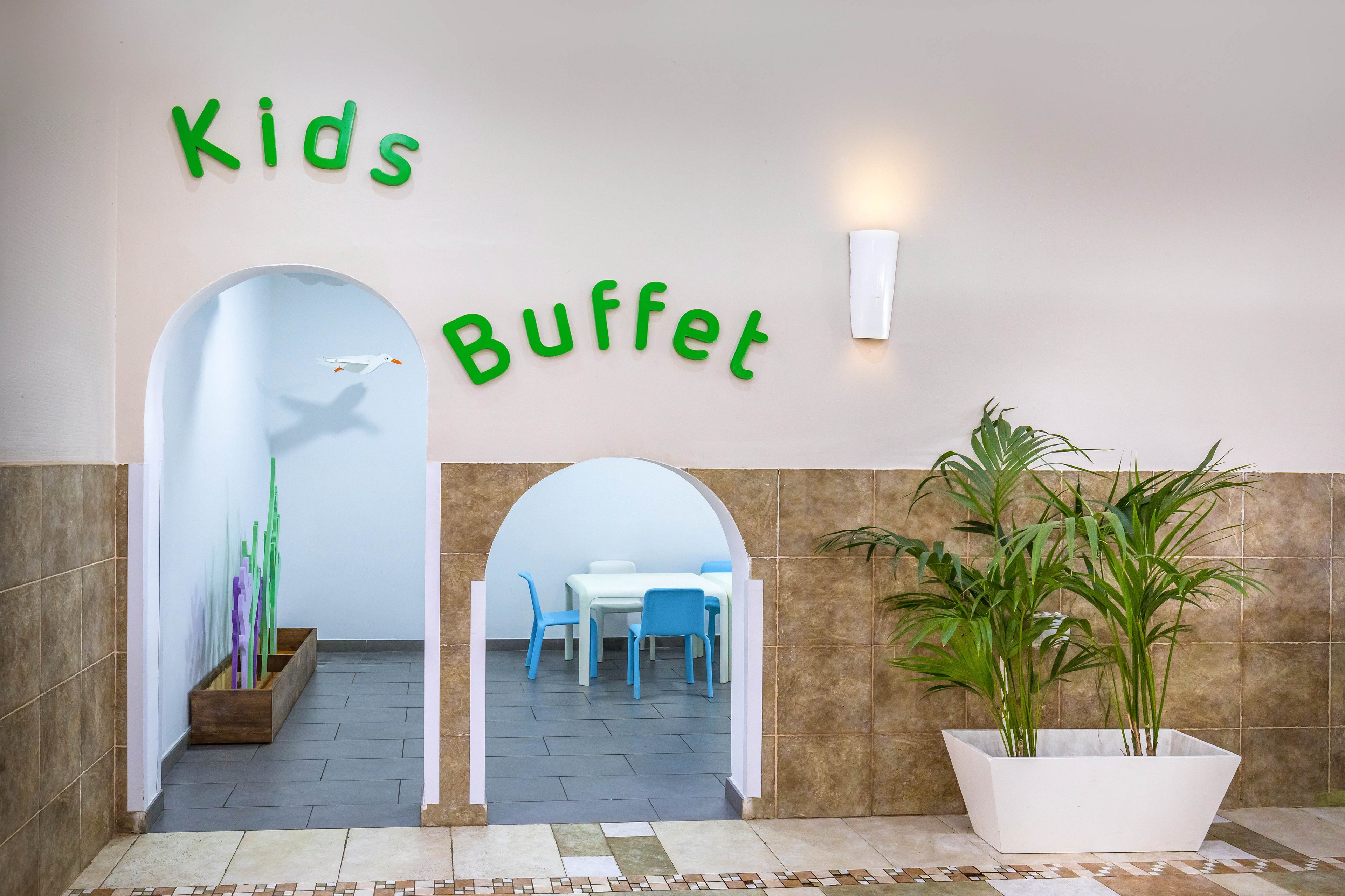 A bright entrance labeled Kids Buffet leads into a child-friendly dining area. Two arched doorways reveal small tables and blue chairs sized for children. The space features tiled walls, a potted plant in a white planter, and playful decor in the background. Soft lighting and clean surfaces create a welcoming, family-oriented atmosphere.